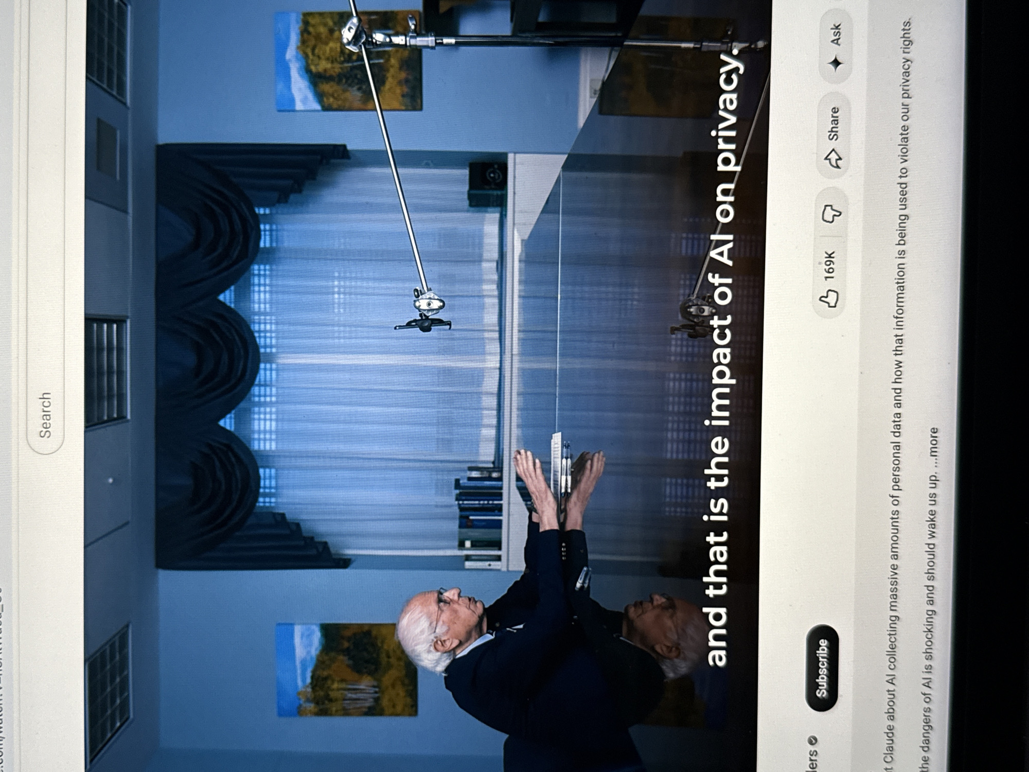 The image shows a video screenshot from YouTube of an interview with an older man with white hair and glasses, wearing a dark blazer. He is standing in front of a window with blinds, with paintings on the wall behind him. There is a boom microphone above him. A reflection of the man is mirrored beside him. Text overlay reads "and that is the impact of AI on privacy." The YouTube interface is visible with the search bar, like/dislike buttons, share button, subscribe button, and a description of the video. The location appears to be indoors, possibly a studio, but the city of New York cannot be determined based on the image alone.