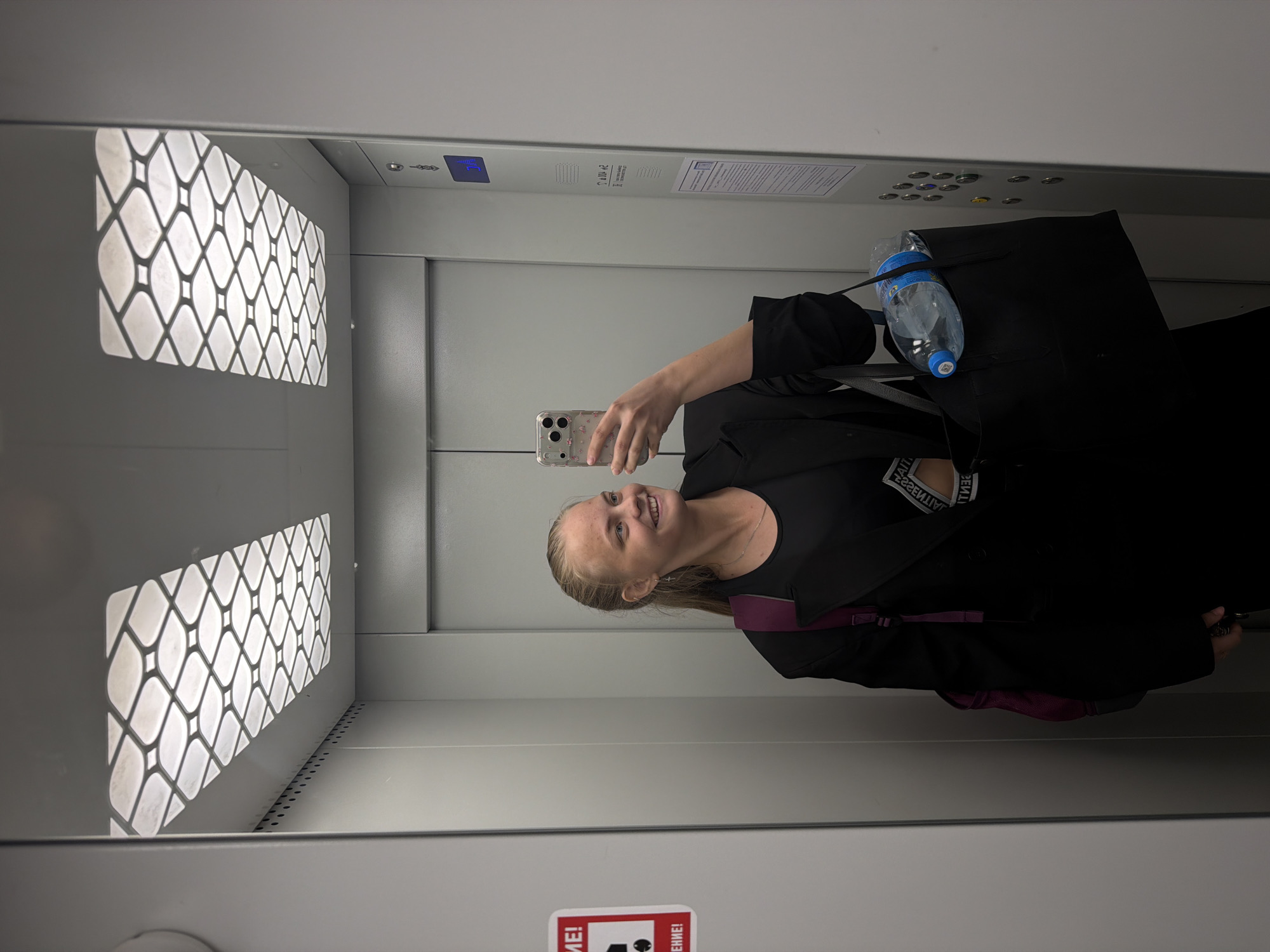 A young woman is taking a selfie in an elevator. She is holding a smartphone in her right hand. She is wearing a black top and has a purple backpack. The elevator has a distinctive grid-patterned ceiling light and various labels and signs in Russian. A water bottle is attached to a black bag. A sign in the lower portion of the elevator is also in Russian. The location appears to be in Russia, possibly Chaikovsky, based on the language on the signs.