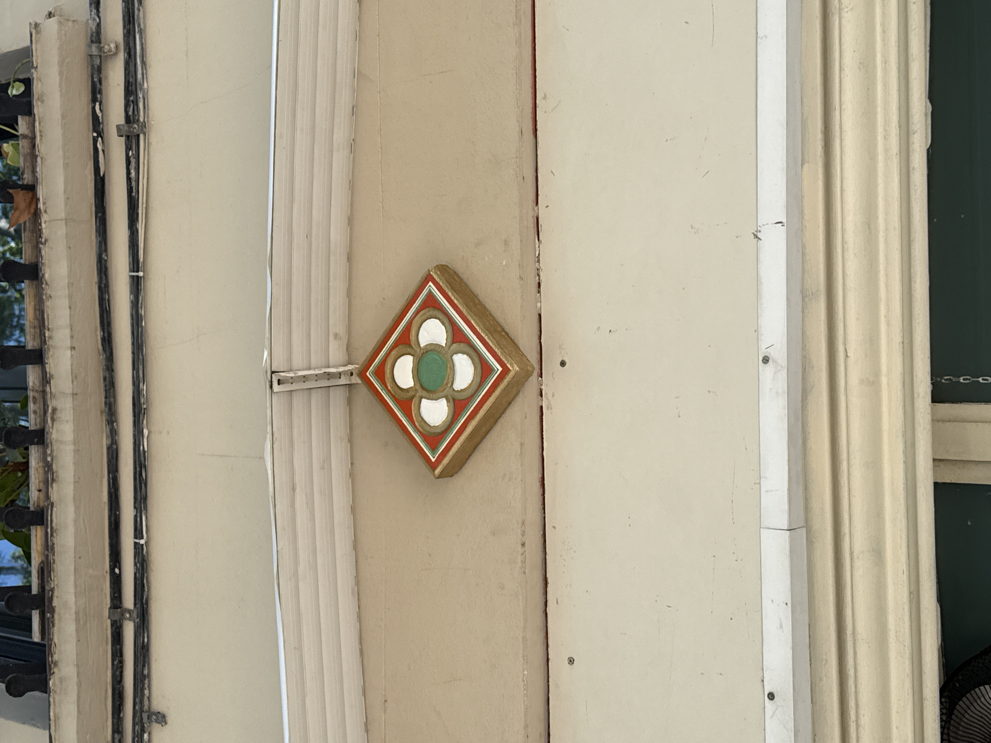 The image shows a portion of a building's exterior. There is a decorative, diamond-shaped, possibly architectural detail featuring a four-leaf clover design with a green center, white petals, red outline, and gold trim. To the left, there is a window and vertical electrical conduits attached to the wall. To the right of the diamond detail, a vertical beam or section is visible. Fluted architecture is also seen on the right side of the frame. Given the architectural details, it is likely located in Barcelona, Spain.