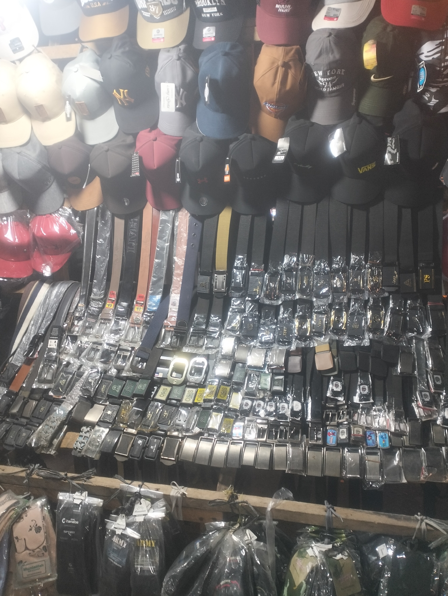The image captures a street market stall in Duri Timur, Indonesia, showcasing a wide assortment of belts and caps. The stall is densely packed with merchandise, with rows of belts hanging vertically and horizontally, their buckles and straps clearly visible. The belts appear to be of various colors and styles, many individually packaged in plastic. Above the belts, numerous caps are displayed, some hanging and others stacked. These caps also come in a variety of colors, designs, and brands, with some featuring logos and text. The overall impression is that of a busy, well-stocked market stall selling accessories.