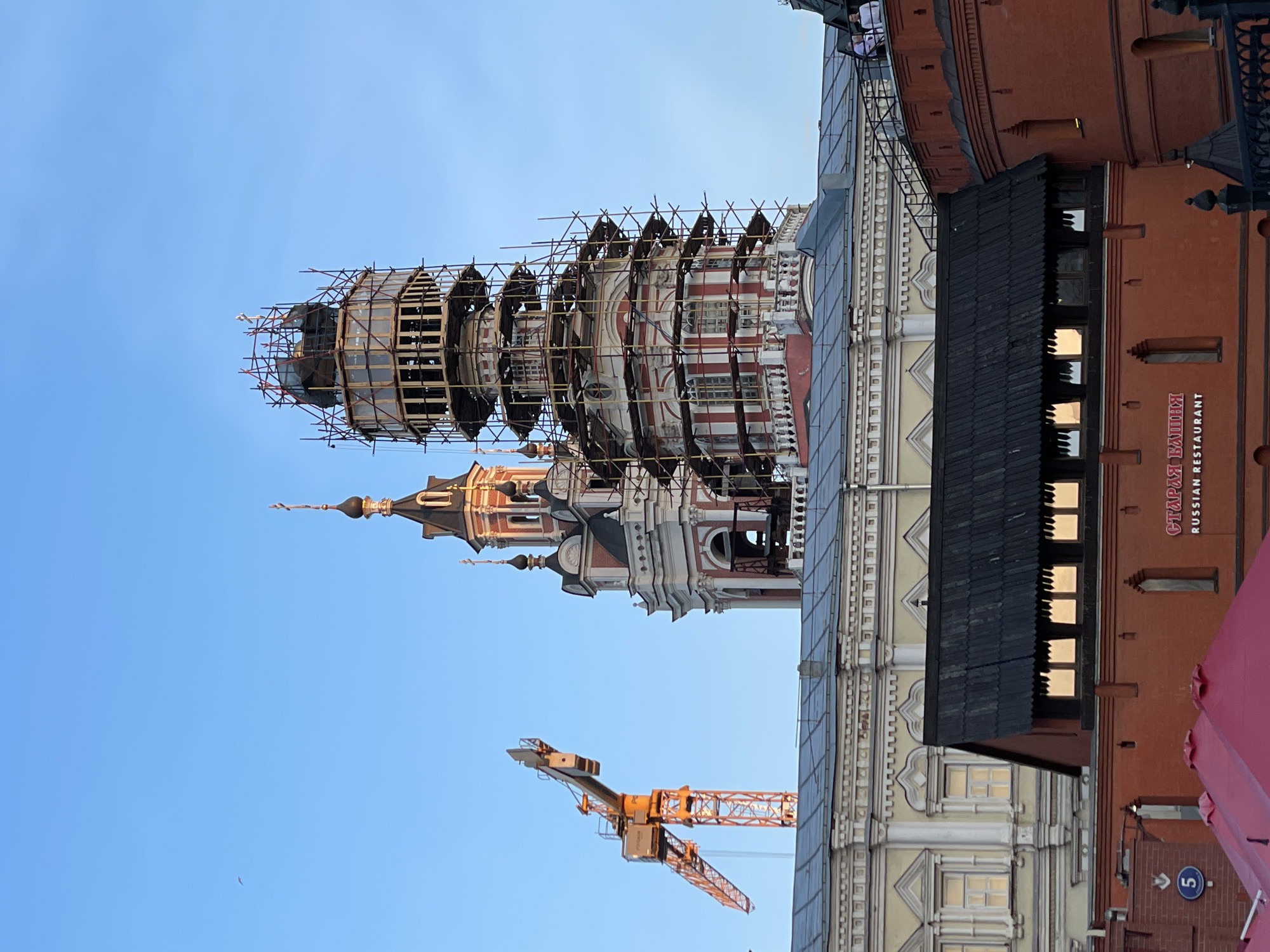 The image depicts a view of the Ivan the Great Bell Tower in the Moscow Kremlin, Russia, undergoing restoration. The tower is partially obscured by scaffolding. A construction crane is visible in the foreground. Adjacent to the tower is a building with a sign indicating a "Russian Restaurant." The sky is clear and blue.