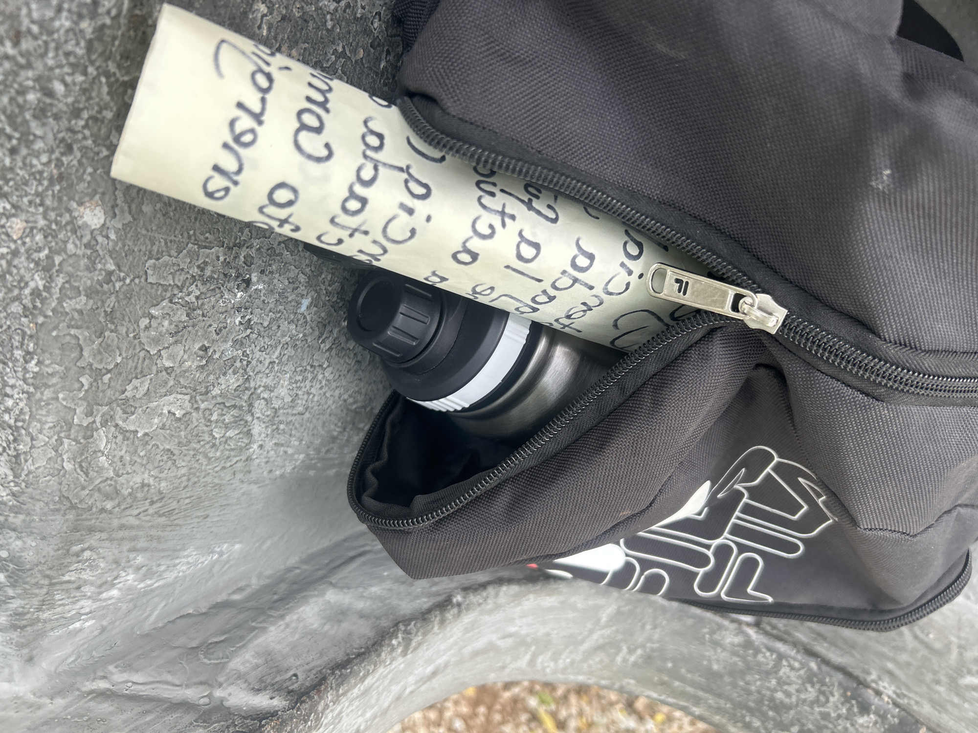 A rolled-up piece of paper with handwritten text in black ink is visible peeking out of the zippered opening of a black Fila brand fanny pack. The fanny pack is partially open, revealing a silver metallic water bottle inside with a black cap. The fanny pack and its contents are resting against a rough, textured gray surface, possibly a concrete bench or a stone structure. The background is out of focus but appears to be an outdoor setting with hints of foliage. The handwritten text on the paper is largely illegible due to the angle and resolution, but some letters and word fragments can be discerned, suggesting it is a note or message. The image appears to be taken in Santa Ana, Venezuela.