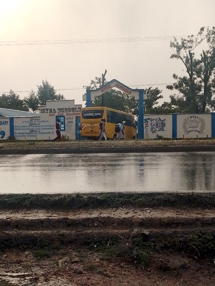 The image shows a yellow school bus parked in front of a school gate. The school gate has an arch with the words "MYRA SCHOOLS DAY AND BOARDING" written above it. There are several people walking by the bus, and the bus is labeled "SCHOOL BUS". The ground is wet, likely from recent rain. The image was taken in front of the Myra Schools Day and Boarding School in Isukha ICHINA, Kenya. The location is confirmed using the provided coordinates (latitude: 0.31, longitude: 34.774). The environment is a school setting with a paved road in front of the school. The weather is overcast and likely rainy.