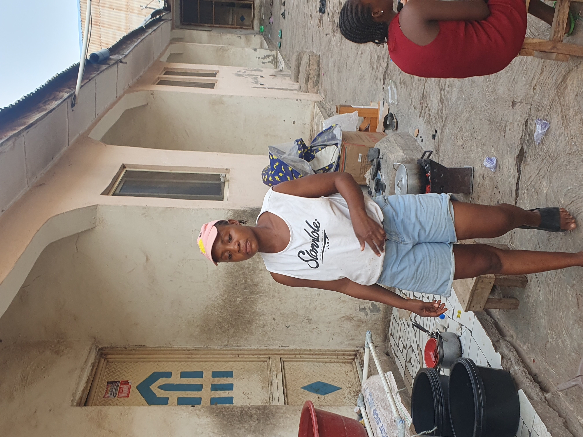 The image depicts an outdoor scene in Lokoja, Nigeria. A young woman wearing a white tank top, denim shorts, and a pink cap is standing in a courtyard with a building behind her. Another person is partially visible, seated on a wooden stool, wearing a red dress. Cooking pots and other items are scattered on the ground. The building appears to be residential, with multiple windows and an aged exterior.