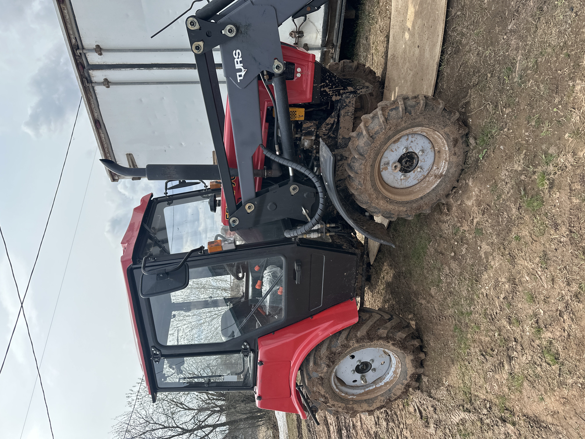 The image depicts a red tractor with a black loader arm labeled "TURS" partially adjacent to a white trailer. The tractor's wheels are muddy, suggesting recent use in wet conditions. The ground is muddy and grassy, indicating an outdoor setting. The image was taken during daylight hours under a partly cloudy sky, near what appears to be trees. The tractor has a cabin, and a license plate is partially visible. There is no verifiable evidence in the image that the location is Ludorvay, Russia.
