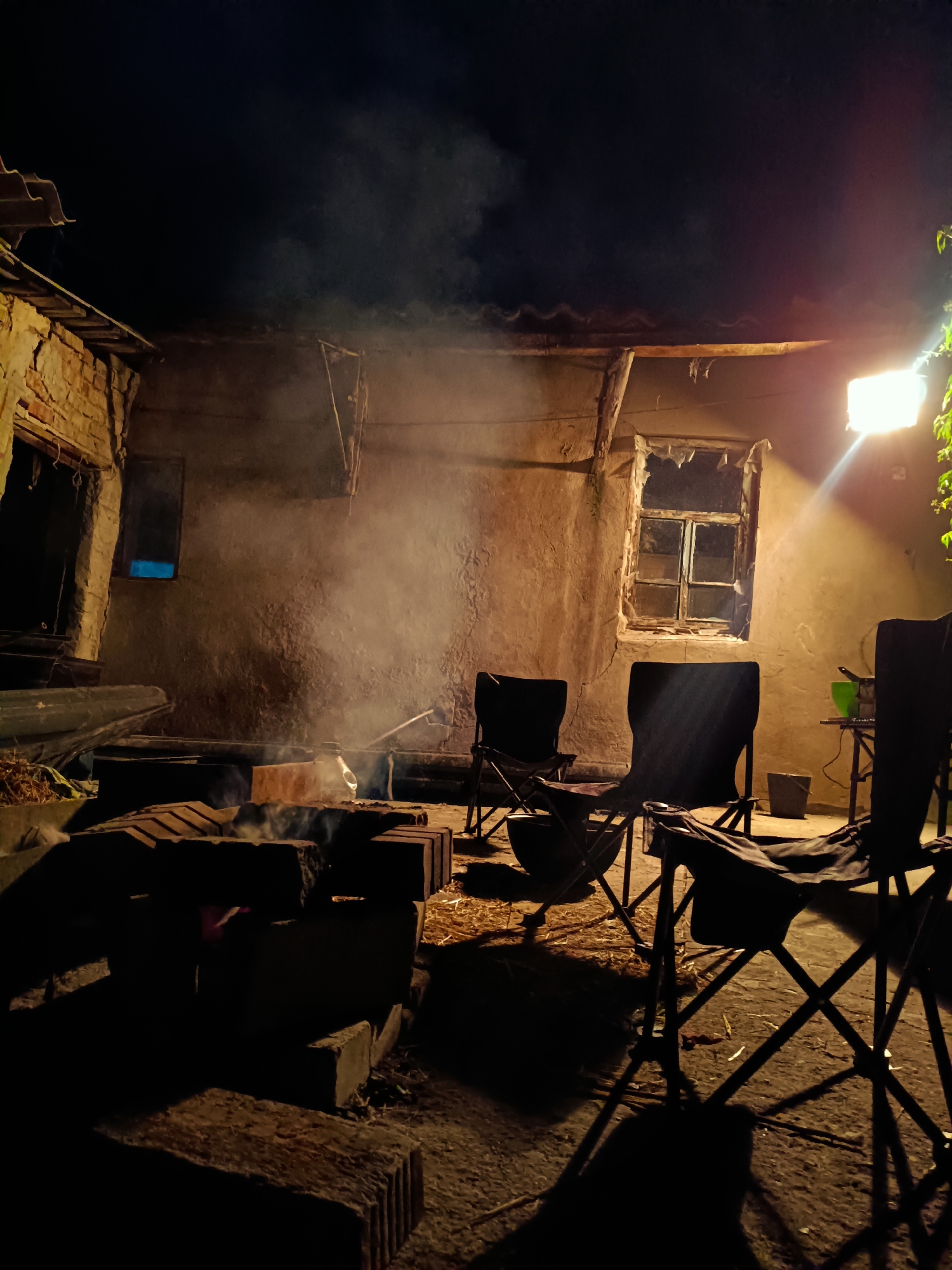 This image captures a dimly lit outdoor scene at night, likely in Tash-Dobo, Kyrgyzstan. The setting appears to be a rustic, somewhat dilapidated courtyard, possibly a backyard or a communal outdoor area. The main subjects are several black folding chairs arranged around what looks like a makeshift fire pit constructed from bricks. Wisps of smoke rise from the fire pit, indicating a fire is either active or has recently been burning.
The surrounding structures are old and weathered, with crumbling walls and a broken window in one of the buildings. A single bright light fixture, possibly a floodlight, illuminates a portion of the wall and casts long shadows across the ground, contributing to the dramatic, nighttime atmosphere. The ground is a mixture of dirt and straw.
The overall mood is one of quiet gathering or anticipation, suggesting a moment of relaxation or camaraderie around a fire. The time of day is clearly night, and there are no immediate indicators of the weather, though the smoke suggests it is not actively raining or windy. The location cues, like the architecture and the presence of outdoor seating around a fire pit, are consistent with a rural or semi-rural setting in Kyrgyzstan. No visible text is present in the image.