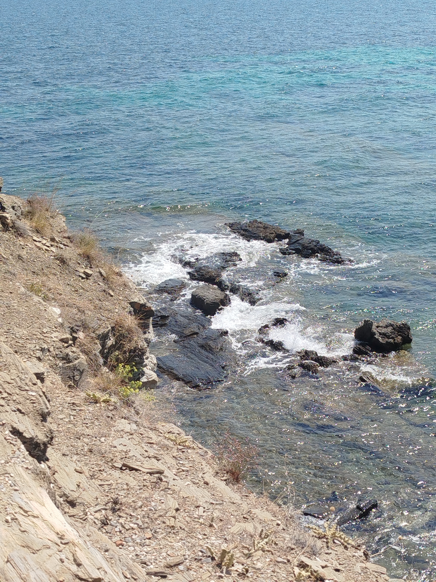 This is a high-angle, full shot of the rocky coastline of Sithonia Municipal Unit, Greece. The image captures the transition from a steep, dry, rocky embankment on the left to the clear, turquoise water of the sea on the right. The embankment is composed of layered sedimentary rock and sparse, dry vegetation. Several dark, jagged rocks protrude from the water near the shore, with waves gently breaking around them, creating white foam. The sea itself is calm, with sunlight glinting off its surface. The scene is uninhabited and devoid of any human presence.