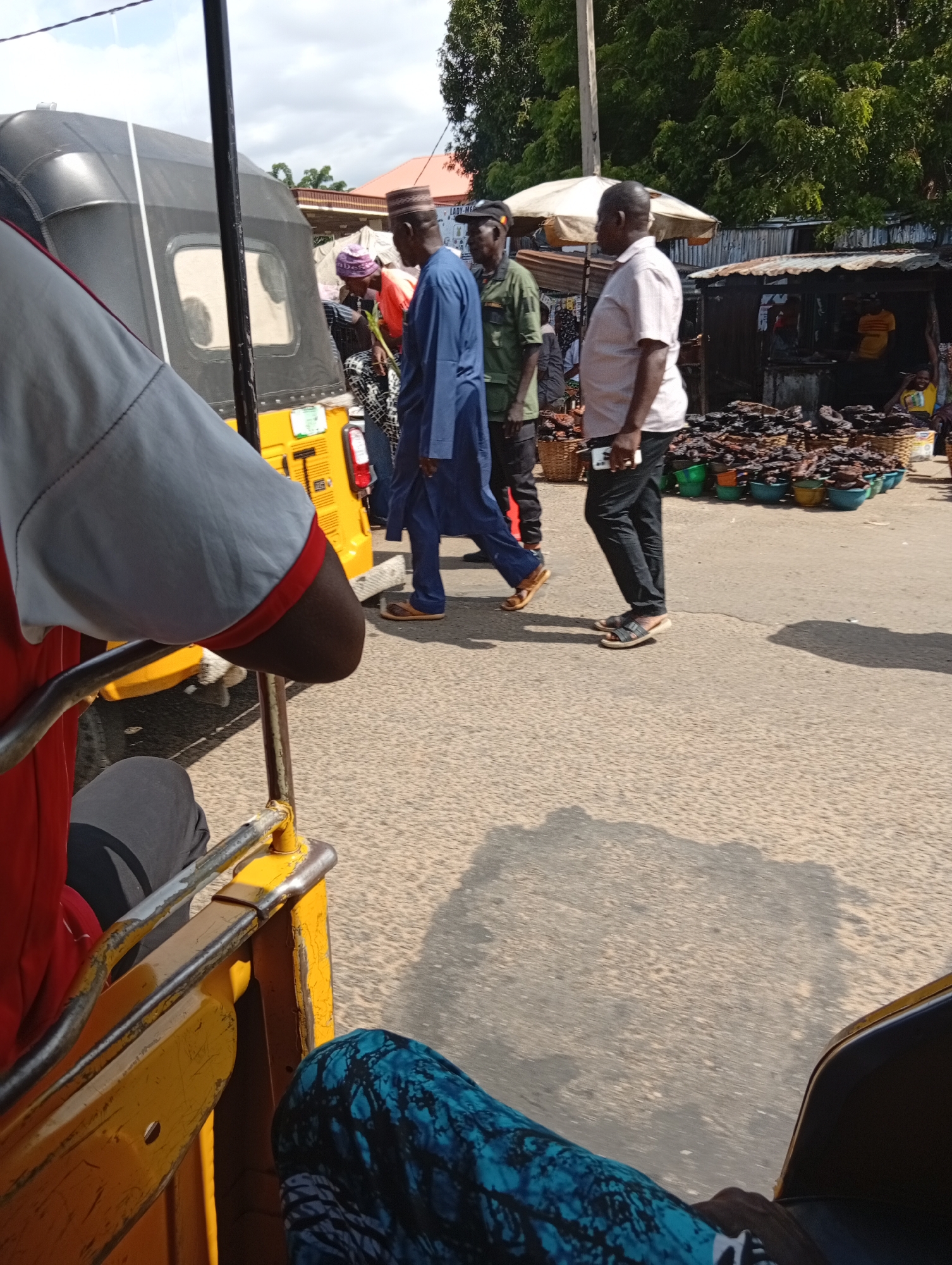 In Jalingo, Nigeria, a street market scene unfolds, viewed from the perspective of someone seated within a yellow auto-rickshaw. In the foreground, the arm of a person wearing a red and grey shirt is visible, resting on the vehicle's railing. Directly ahead, a black-covered auto-rickshaw is parked. Several individuals are gathered near the auto-rickshaw.
One man, wearing a blue traditional outfit and a cap, walks away from the camera. Another man in a green shirt and cap stands beside him, facing the auto-rickshaw. Further into the scene, a man in a light pink shirt and dark pants is looking towards the market stalls.
The market itself features several stalls, with awnings providing shade. Piles of dried fish are displayed in baskets and bowls. Behind the market stalls, trees and buildings are visible. The ground is paved, and shadows from the structures and people are cast across it. The sky is partly cloudy.