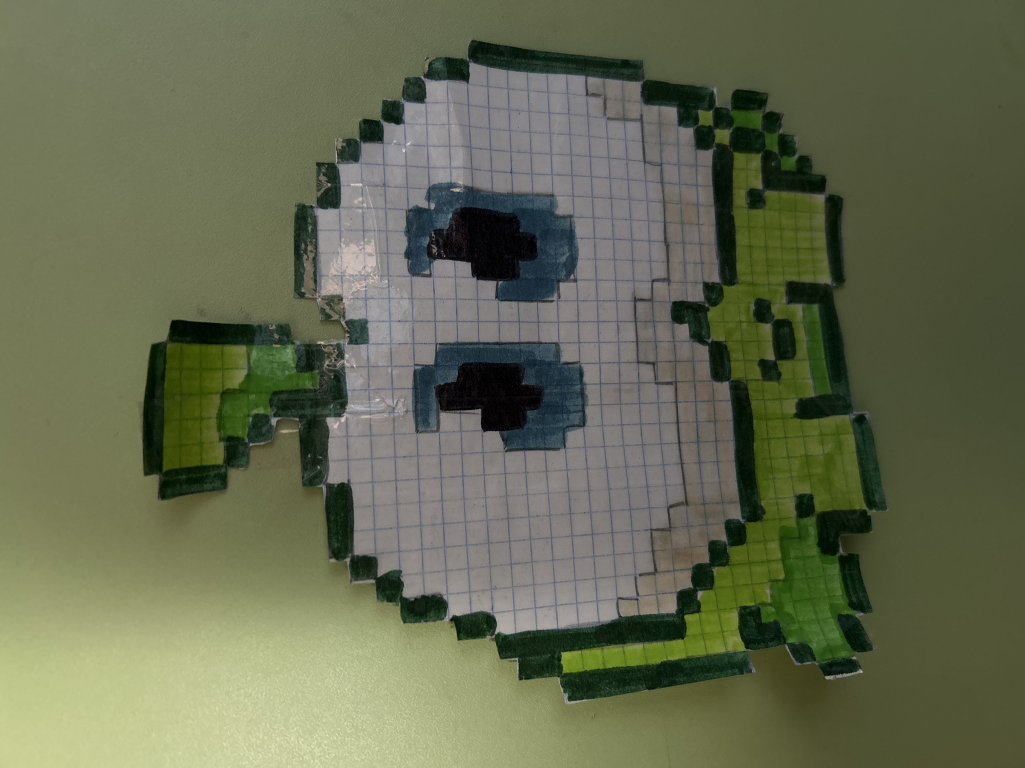 An image displays a hand-drawn pixel art cutout placed on a smooth, light olive-green surface, likely a desk or table, in Kerch, Ukraine. The main subject is a character resembling a cartoon ghost or an abstract creature, rendered in a pixelated style on graph paper.
The character has a predominantly white, roughly oval-shaped body with visible graph paper lines underneath the coloring. It features two prominent, rectangular black eyes, each outlined with a lighter blue color. Extending from the character's left side (viewer's right) is a large, vibrant green, leaf-like or arm-like appendage, also rendered in pixels with a darker green outline. A smaller, similar green pixelated element is visible on the character's right side (viewer's left). The green coloring varies in shade, suggesting different tones were used or varying pressure during coloring.
The entire drawing is cut out along its pixelated edges, with some slight irregularities indicating a handmade cut. Portions of the cutout appear to be slightly raised from the surface. The background is uniform and featureless, emphasizing the artwork. No specific activity is taking place beyond the static display of the artwork. There is no visible text in the image. The lighting suggests an indoor setting, likely under artificial light.