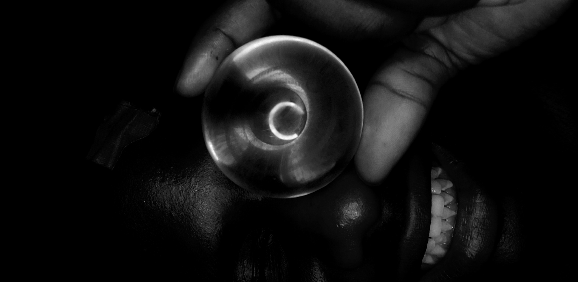 A black and white photograph shows a person’s face with a hand holding a translucent, hollow sphere over their nose. The person’s mouth is partially open, revealing their teeth. A small, dark, rectangular object is visible near the person’s forehead. The photograph is dimly lit, with most of the scene obscured in darkness. The location is not determinable from the image.