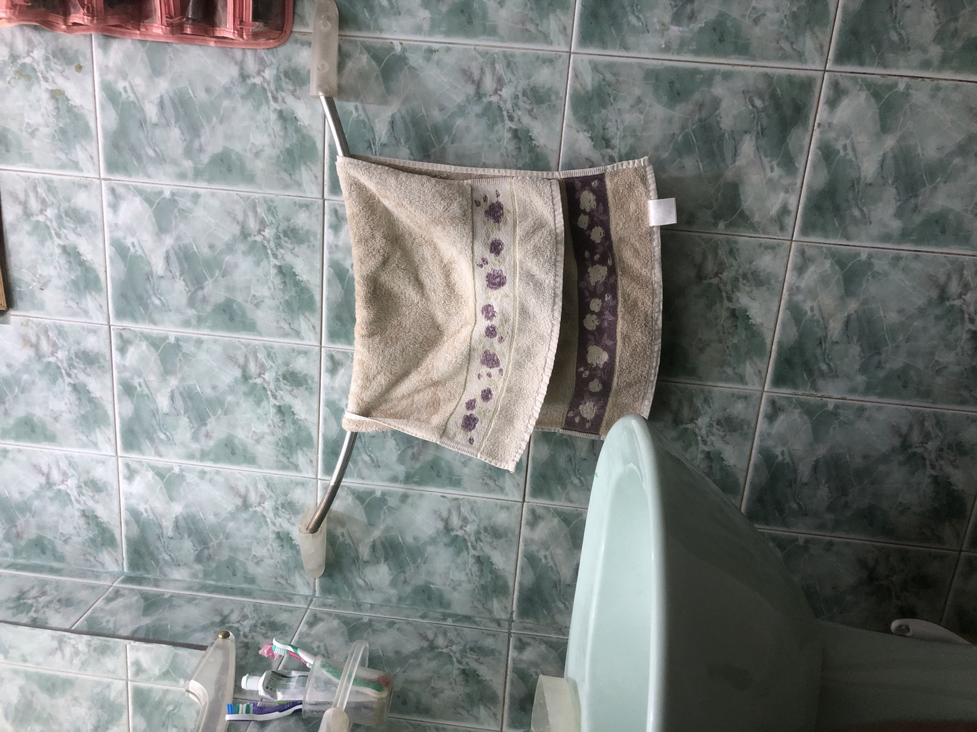 The image depicts a close-up view of a bathroom sink and a towel rack mounted on a tiled wall. The sink is a light blue ceramic basin, and the wall is covered in square green marble-patterned tiles. A metal towel rack is installed horizontally on the wall, holding two folded towels. The top towel is a light tan color with a patterned border featuring small floral designs in purple. The bottom towel is also tan and has a similar patterned border. To the left of the towel rack, a clear plastic holder contains toothbrushes and a small bottle of what appears to be hand sanitizer. The lighting suggests an indoor environment, likely a residential bathroom. The scene is static and shows no people or ongoing activities.