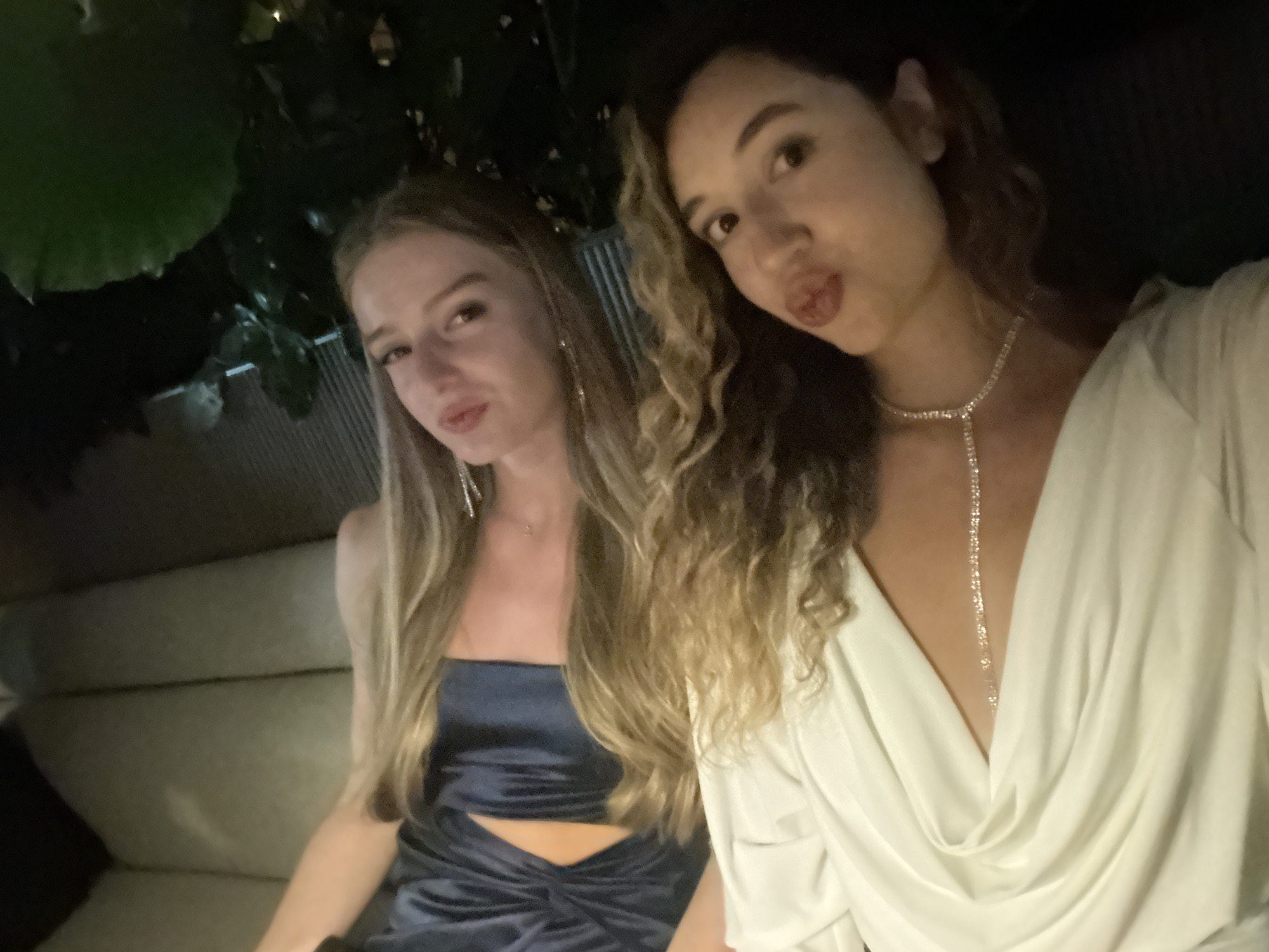 Two women are seated on a sofa, posing for a selfie. The woman on the left has blonde hair and is wearing a dark blue dress. The woman on the right has curly brown hair and is wearing a white dress with a sparkly necklace. They are in a dimly lit indoor environment, possibly a restaurant or lounge, with plants visible in the background. The image appears to be taken at night.