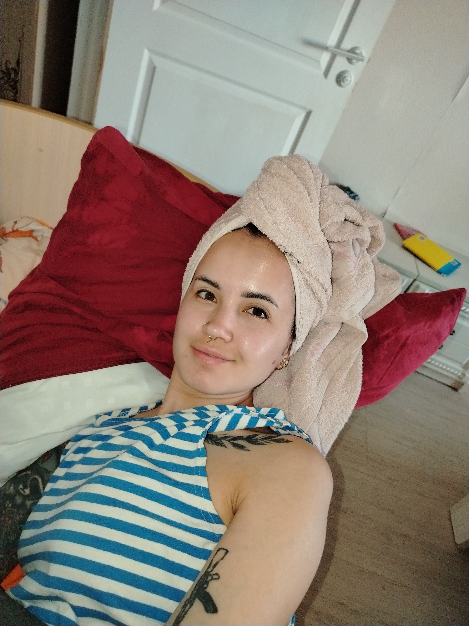 A young woman is lying on a bed. She has a towel wrapped around her head. She is wearing a blue and white striped tank top. There are tattoos on her arms. She is smiling. The bed has red pillows and a white sheet with a patterned blanket. There is a white door and a wooden floor in the background. There is a yellow box near the top right of the frame. The location is not discernible from the image.