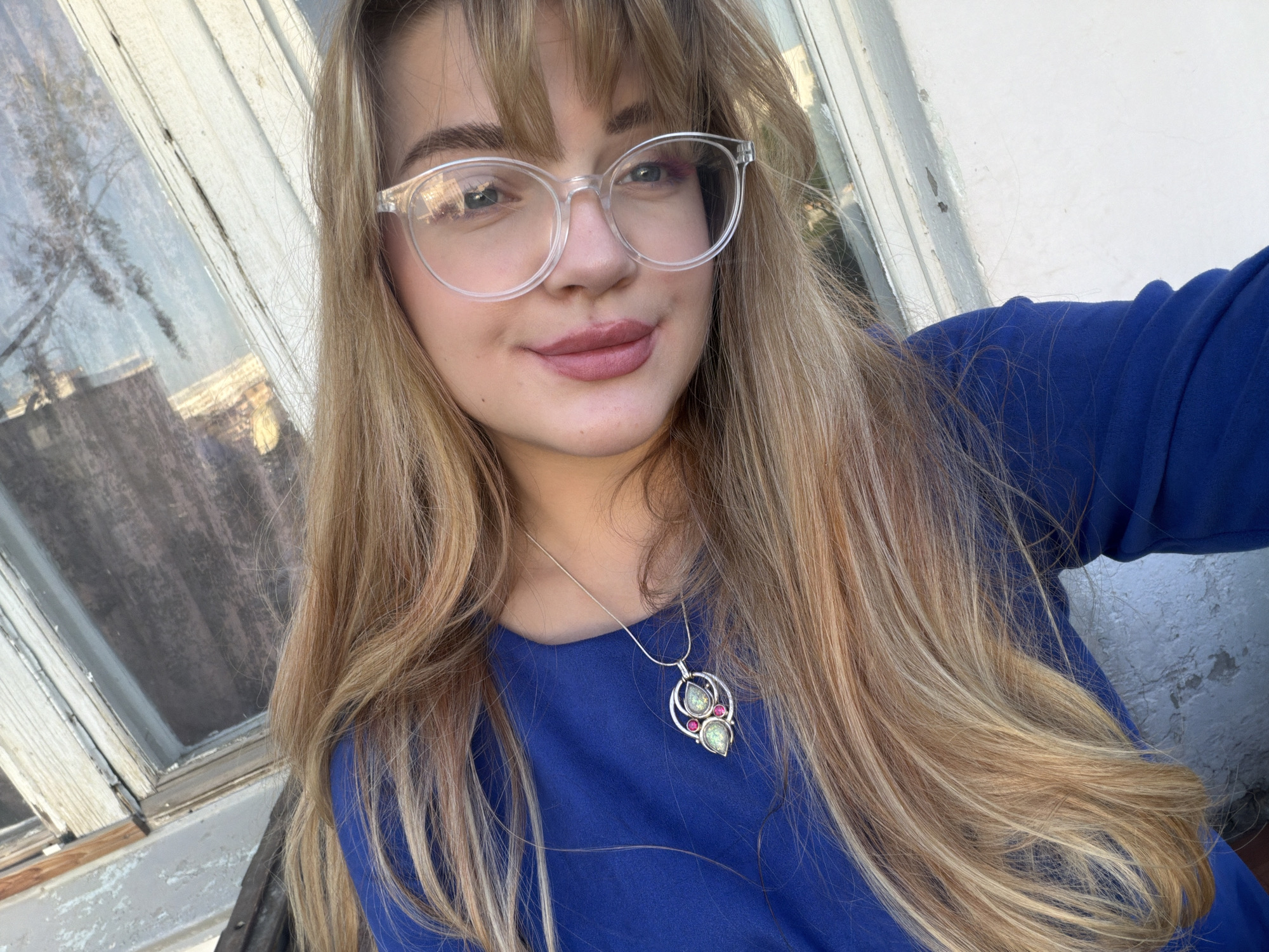 A fair-skinned young woman with blonde hair and bangs poses for a selfie. She wears clear-framed glasses and a blue top. A silver necklace with a pendant is visible. The background includes a window with a white frame and possibly a view of a building or cityscape. The location is Naberezhnye Chelny, Russia.
