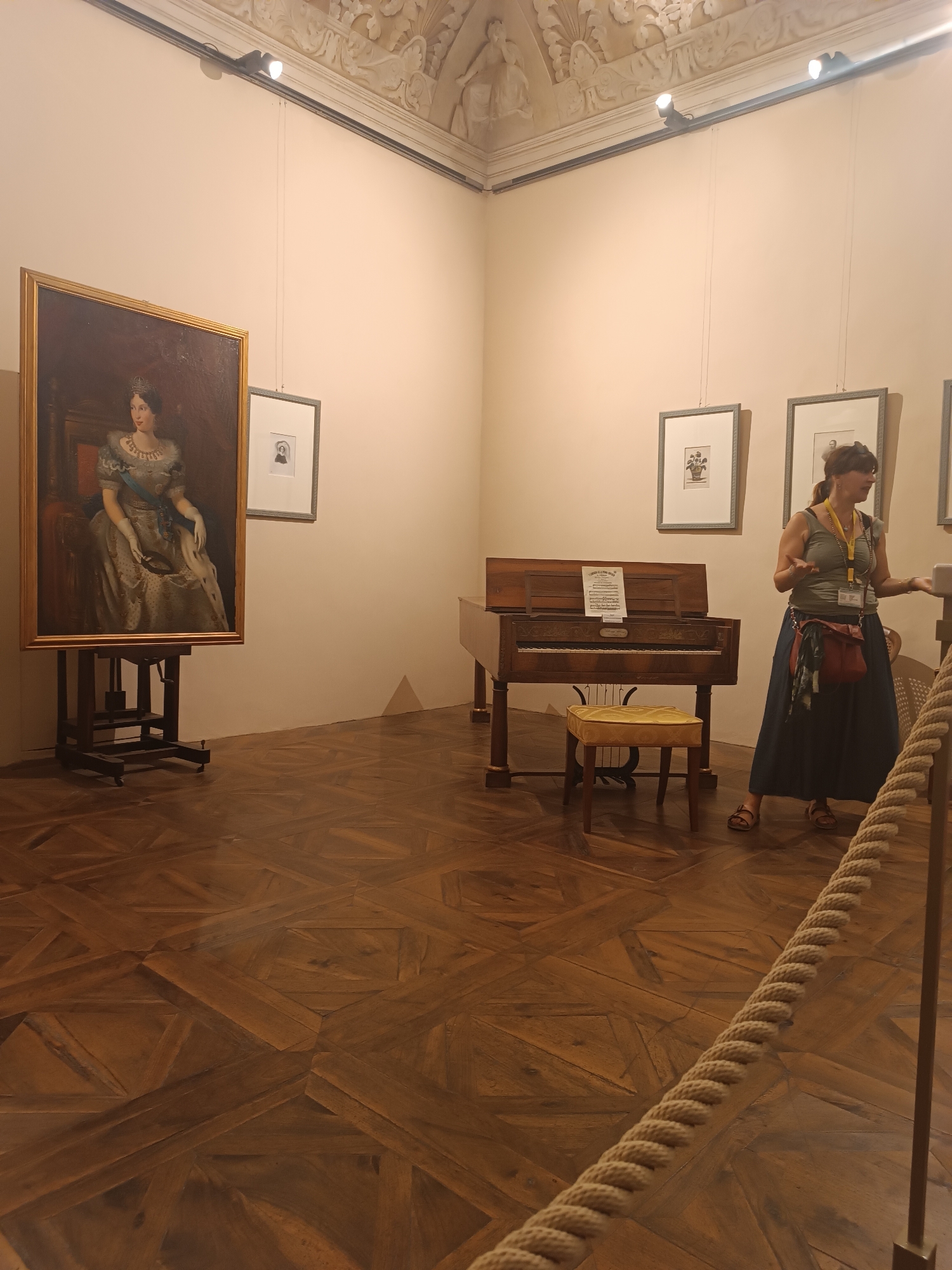 In a museum gallery in Colorno, Italy, a woman stands near a historic piano, gesturing as if explaining something. She is wearing a green tank top, a long navy skirt, and sandals. A red purse hangs from her shoulder, and she has a lanyard with badges around her neck. The room features a richly patterned wooden floor and a high ceiling with ornate plasterwork.
To the left of the piano, a large framed portrait of a woman in a formal gown and crown is displayed on an easel. Next to the portrait, two framed prints hang on the wall. The piano itself is an antique instrument with sheet music on its stand and a small yellow stool in front of it. The lighting in the room is provided by spotlights mounted on the ceiling. The scene appears to be set during the daytime, as indicated by the ambient light and artificial lighting. The overall atmosphere suggests a guided tour or educational presentation within the museum.