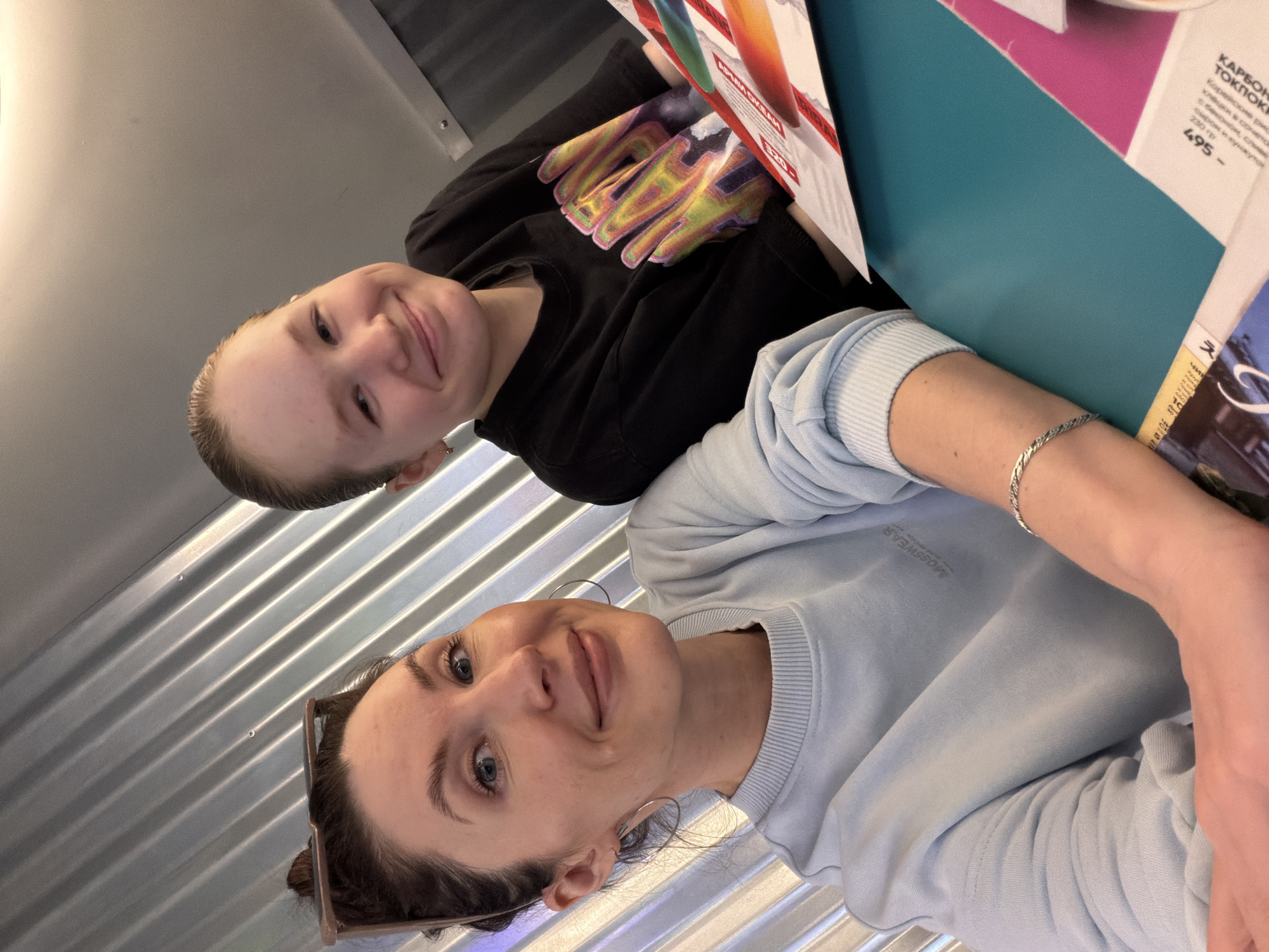 Two people are positioned in a photo taken from a high angle. The person in the foreground is wearing a light blue sweater and silver hoop earrings, with her dark hair pulled back. A silver bracelet is visible on her wrist. Behind her, another person wears a black sweater with a colorful hand design. They are sitting at a table with menus and promotional materials. One menu includes a section in Russian for a dish called "CARBON TOKP" and the price of 495 rubles. The location appears to be in Perm, Russia, based on the Russian text. A corrugated metal wall is visible in the background.