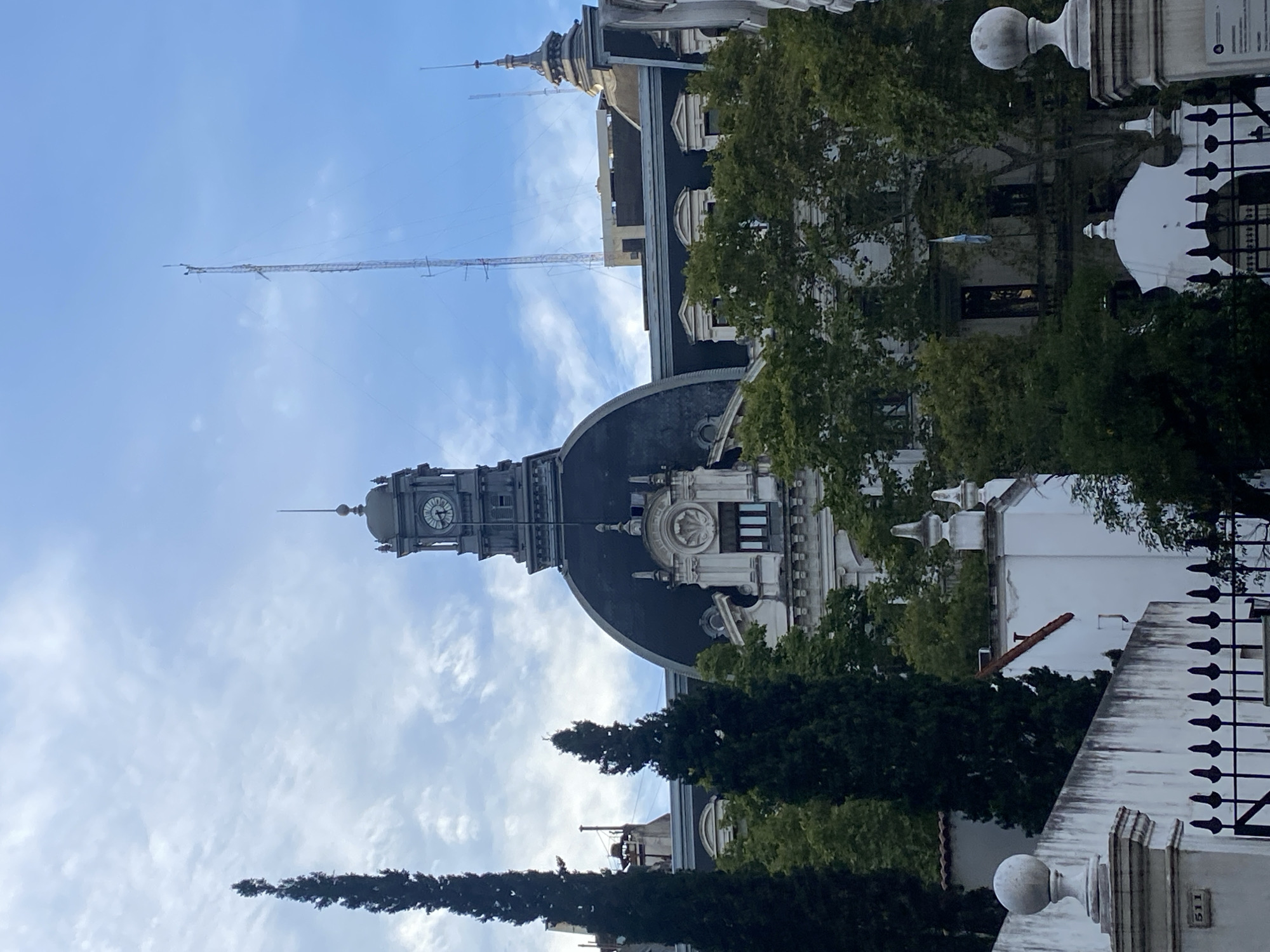 The image shows a view of the Palacio de Gobierno de la Ciudad de Buenos Aires, located in the city of Buenos Aires, Argentina. The view is from a low angle, capturing the building's facade, dome, and clock tower against a cloudy, blue sky. Several antennae rise from the building's top. Trees partially obscure the view of the building. A fence and gate are visible in the foreground.