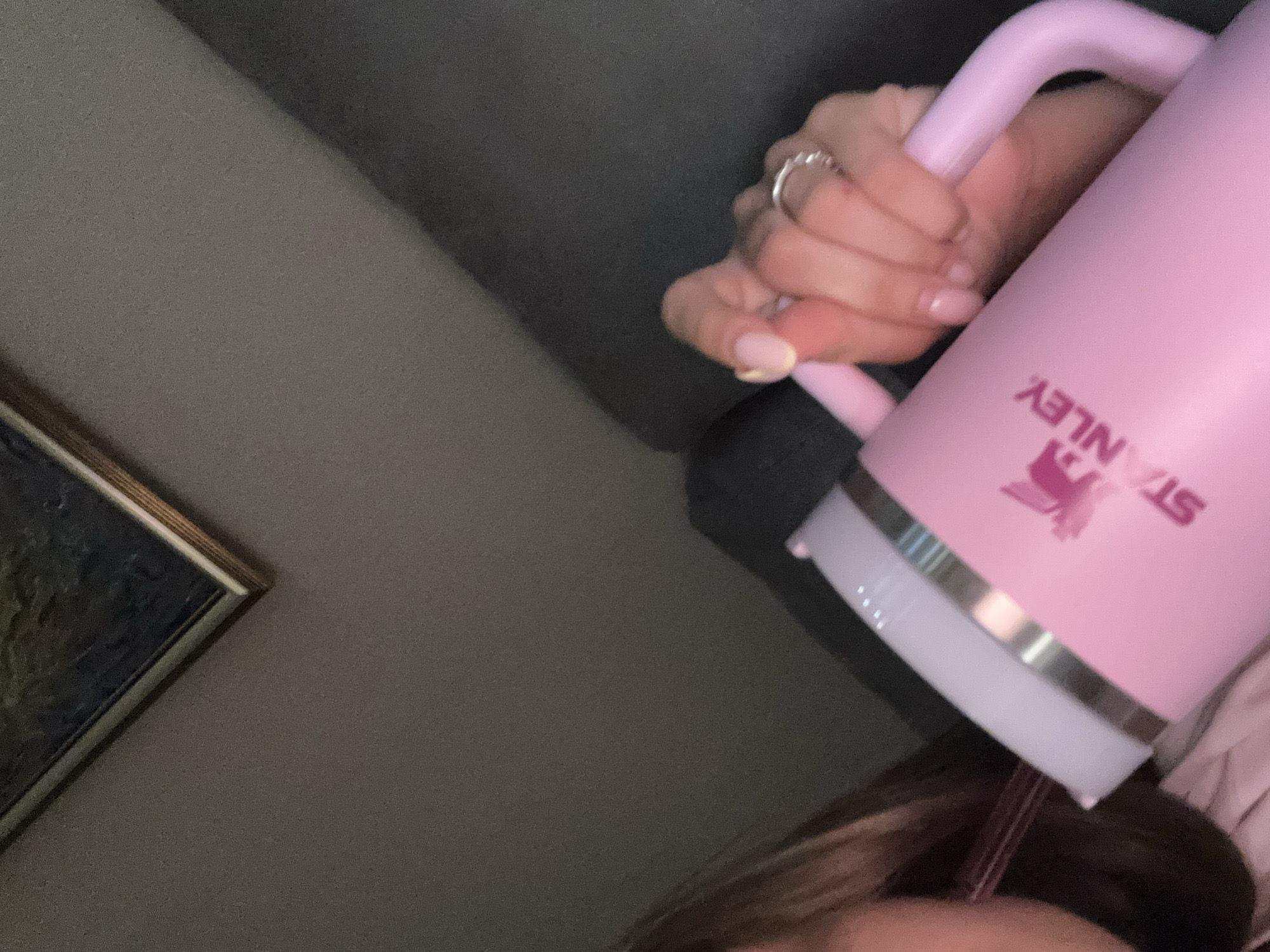 A close-up, low-angle indoor shot shows a person's hand holding a pink Stanley tumbler with a white lid and a pink straw. The tumbler has a pink logo and the word "STANLEY" printed in pink. The person is wearing a ring on their left hand. The background consists of a dark gray couch and a framed picture on a wall. The scene is likely in a residential setting in Sofia, Bulgaria.