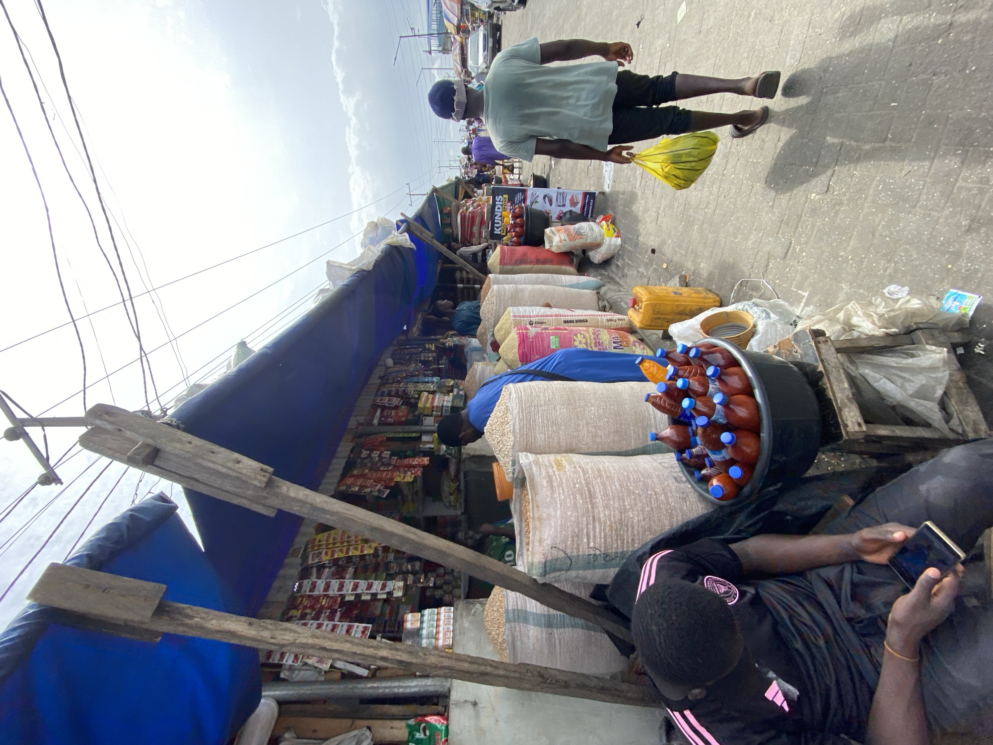 The image captures a street scene in Lekki, Nigeria. A person wearing a light green shirt and black shorts walks to the right, holding a yellow plastic bag. They are walking on a paved area that shows signs of wear.
A roadside market stall dominates the left side of the frame. The stall is constructed with wooden supports and covered with a blue tarp. Various products are displayed, including stacks of goods in bags, packaged snacks, and bottles of what appears to be palm oil, stored in a black container.
A man is seated on the ground next to the market stall, looking at a mobile phone. The man is wearing a black jacket with pink stripes. The area is filled with goods and some discarded items, indicating a bustling market environment. Electrical wires are visible overhead, crisscrossing the sky.