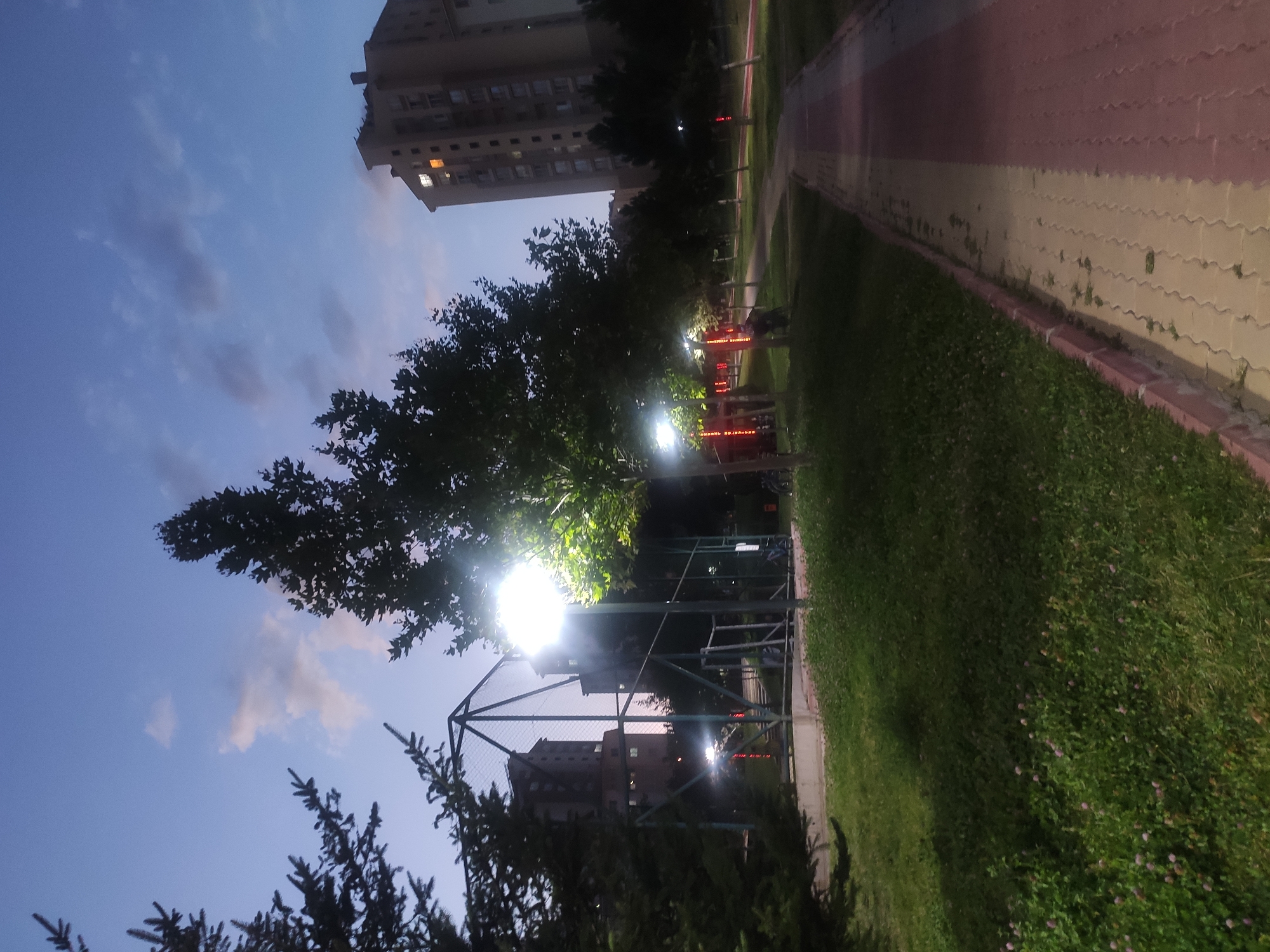 The image depicts a park area in Etimesgut, Turkey, at dusk. A paved walkway with alternating light yellow and red brick sections runs diagonally from the lower right towards the middle ground. To the left of the walkway is a grassy area with scattered small pink flowers. Several trees with green foliage are interspersed throughout the park, some illuminated by streetlights. In the background, there are apartment buildings, one of which has a few illuminated windows. A metal sports court structure is visible to the left of the frame. The sky is a deep blue with some wispy clouds, indicating it is late evening.