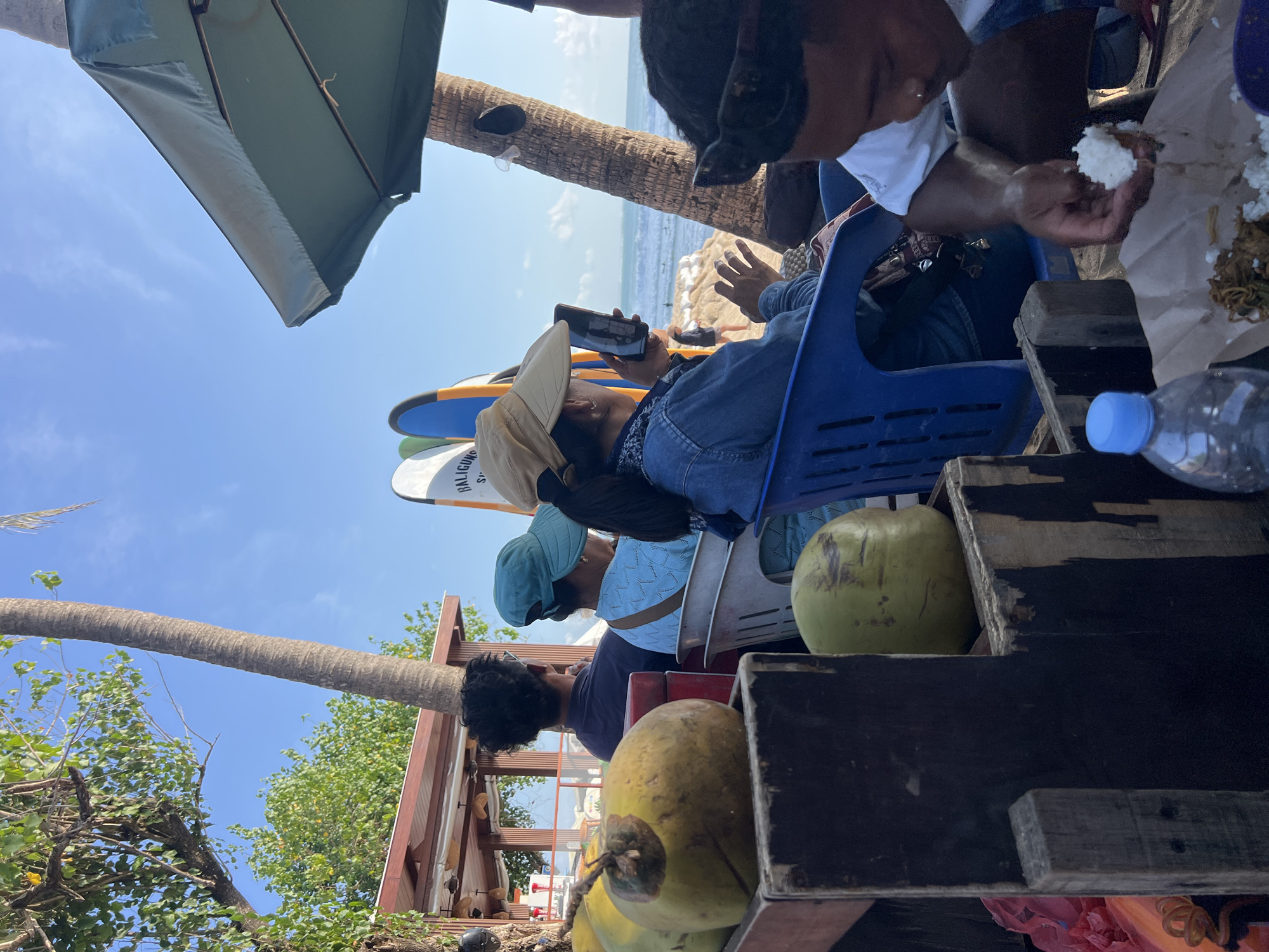 In Kuta, Indonesia, this image captures a scene at the beach with several people visible. In the foreground, there are coconuts in wooden crates. Two people are seated, facing away from the camera, with a woman in the center holding a smartphone and taking a picture. Behind them, surfboards are propped up. To the right, a man is eating from a plate of food. The background shows the ocean with surfers in the water and a blue sky with scattered clouds. The environment suggests a sunny day at a popular tourist destination.