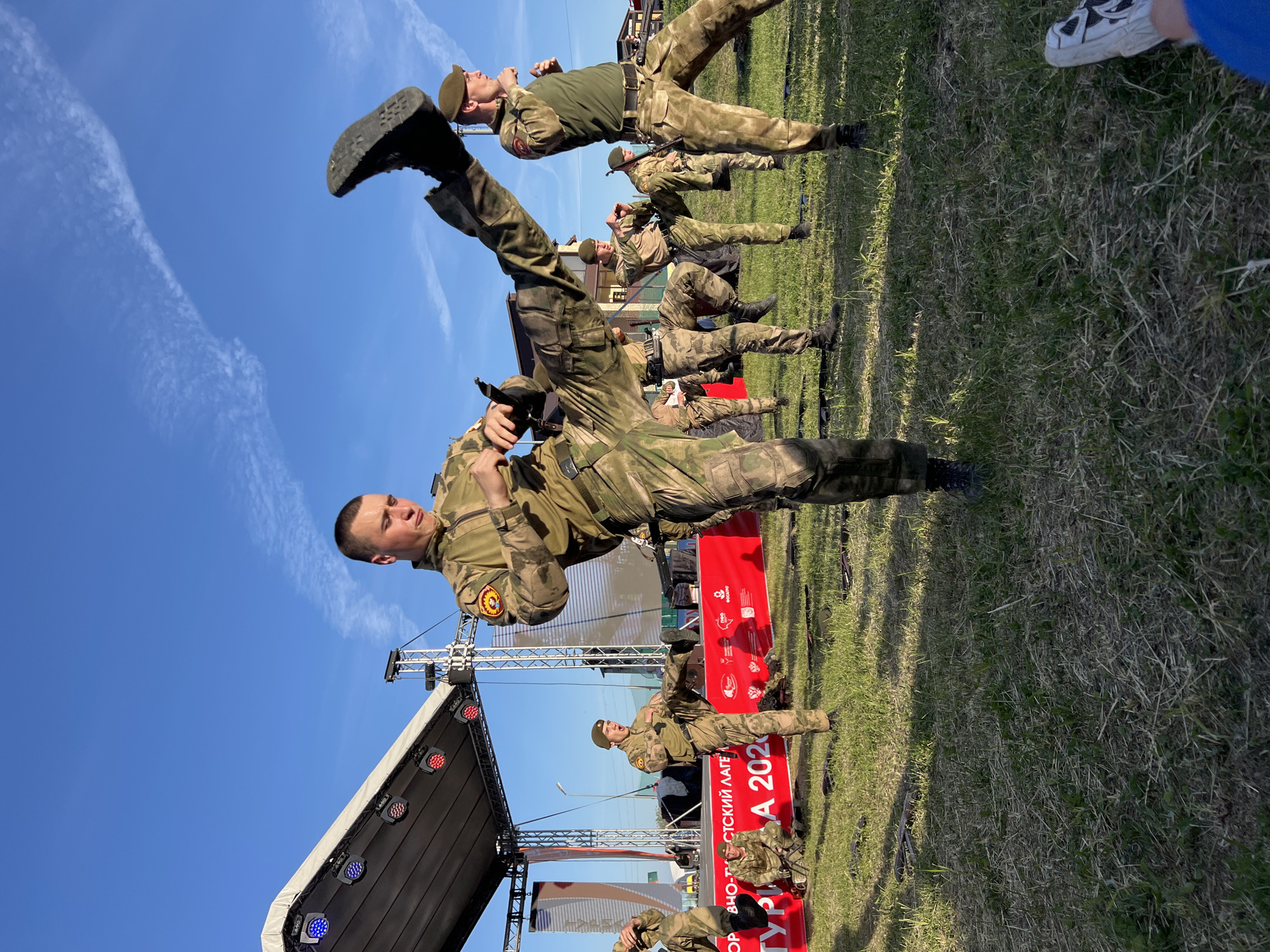 The image shows a military performance in Khvalynsk, Russia. A group of soldiers in camouflage uniforms are performing martial arts moves on a grassy field in front of a stage. The sky is blue and sunny. Some of the soldiers are holding weapons. A red banner with Russian text is visible. Spectators are present.