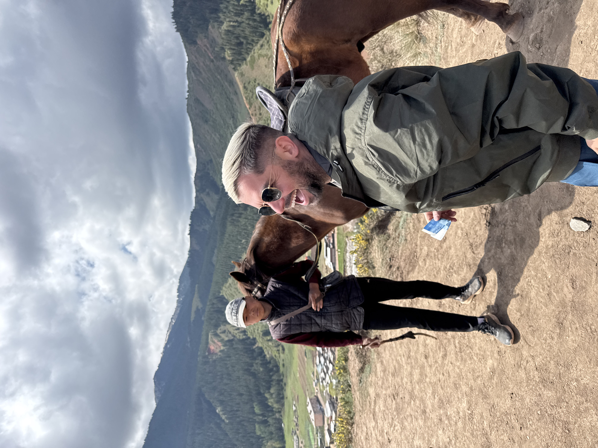 This outdoor image, taken in Jeti-Ögüz, Kyrgyzstan, is rotated 90 degrees clockwise, depicting a mountainous landscape under a cloudy sky.
In the foreground, a white man with blonde hair, a beard, and sunglasses is captured mid-laugh or shout, his mouth wide open in an expression of clear joy. He wears a dark olive green jacket and holds a crumpled blue paper with faint white text in his left hand. The head of a brown horse is positioned close to his face, suggesting interaction, possibly feeding or playful engagement.
Standing behind and to the left of the main subject is another person, appearing to be an Asian male. He wears a white cap with the word "VINTAGE" clearly visible, a dark sleeveless puffer vest over a burgundy long-sleeved shirt, and dark pants. He holds a stick or whip in his right hand and is positioned next to another brown horse. His expression is neutral.
The ground beneath the subjects is dry, dusty earth. The background showcases vast, tree-covered mountains that define the rugged terrain. A valley with a small village, characterized by scattered buildings with light-colored roofs, is visible nestled among the hills. The sky is overcast with a mix of grey clouds, indicating daytime conditions without direct sunlight. The scene suggests a pleasant horse riding excursion or tour in this scenic, rural mountainous region.