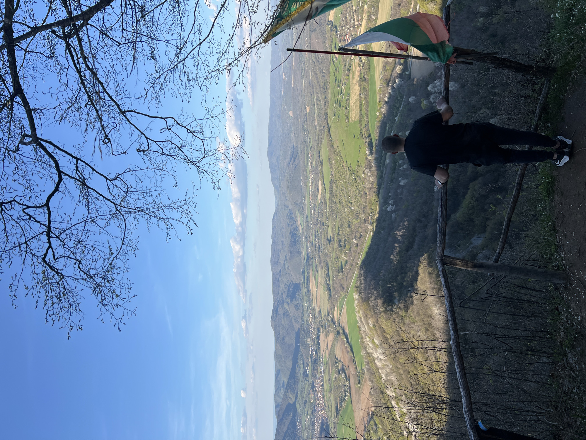 A man in black clothing and sneakers is leaning on a wooden fence at the edge of a cliff, overlooking a valley with fields and trees. A flag with green, white, and red stripes is attached to a pole near the fence. The sky is blue with some clouds, and a tree branch with bare branches is visible in the top left corner. This scene is located in the city of Elena, Bulgaria.