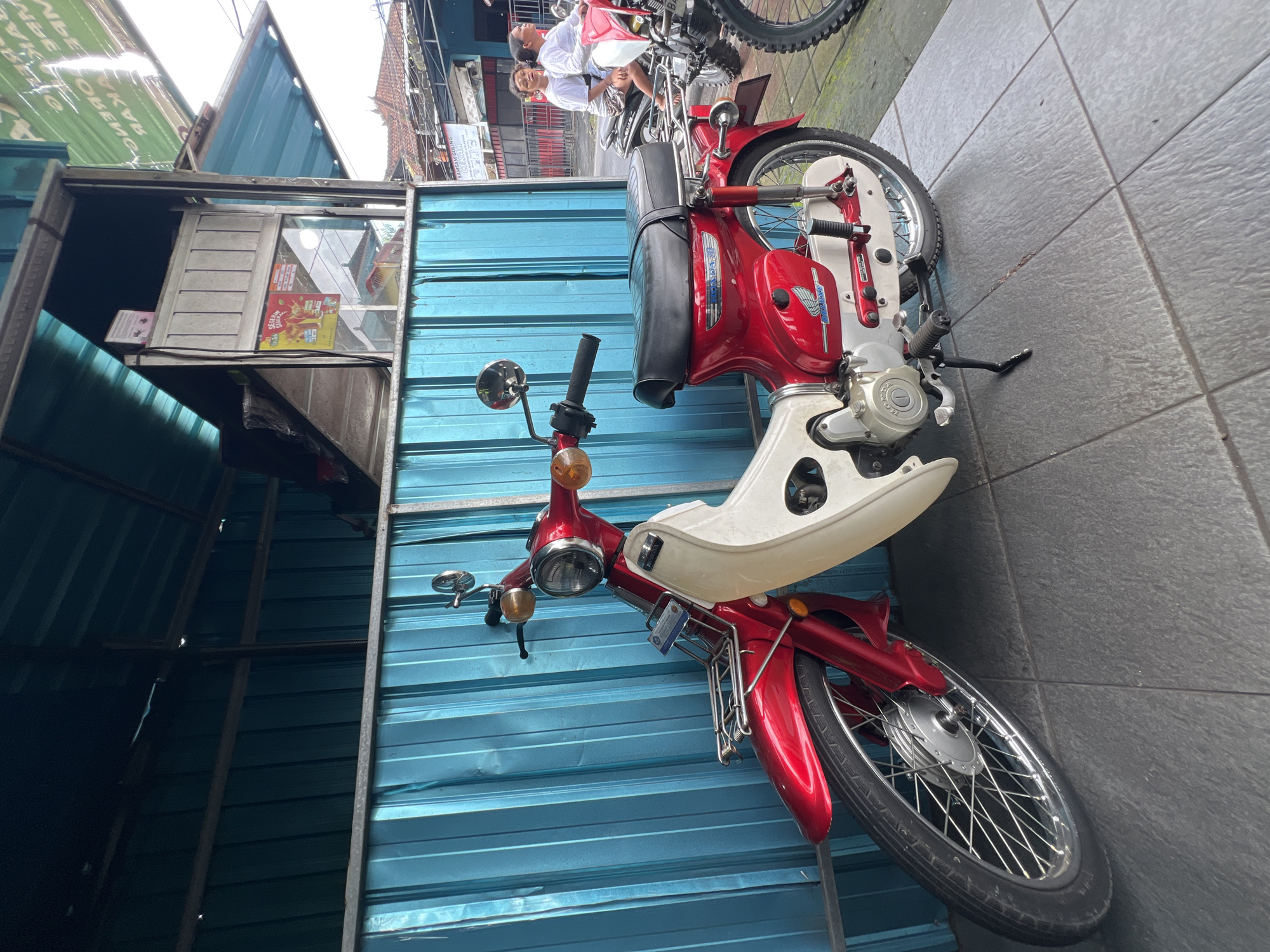 A red and white Honda C70 motorcycle is parked on a tiled sidewalk in front of a blue corrugated metal wall. The motorcycle is facing left and is propped up on its kickstand. It has a chrome front rack, a black seat, and a silver engine. In the background, two people are riding on another motorcycle. To the right of the image, a sign for "SAMSAM GULING" (suckling pig) is visible. The location is indicated to be Gianyar, Indonesia. The area appears to be an urban or semi-urban setting with commercial establishments.