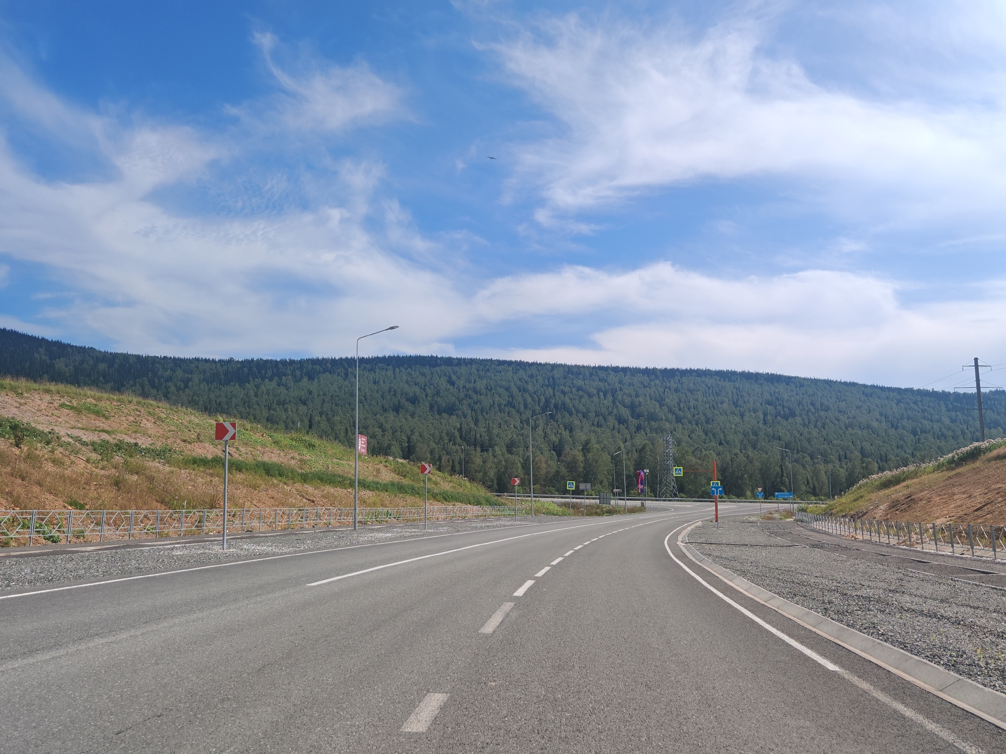 The image shows a paved road curving to the right, with two lanes in each direction separated by a dashed white line. The road is bordered by a gravel shoulder and a metal fence on the right side. On the left side, there is a grassy embankment with some sparse vegetation and a metal guardrail. Beyond the embankment, a dense forest covers a hillside that stretches across the horizon. The sky is a bright blue with scattered white clouds. In the distance, there is a power line tower and a few streetlights. Traffic signs are visible along the roadside, including a directional arrow sign and a pedestrian crossing sign. The scene appears to be in a rural or semi-rural area, possibly on the outskirts of the city of Sheregesh, Russia, given the presence of a forest and the mountainous terrain. There are no people or vehicles visible in the image.