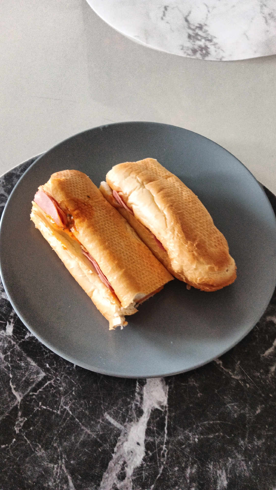 Two ham and cheese subs are arranged on a gray plate, which rests on a black and white marble countertop. The subs are made with long, crusty bread rolls, cut lengthwise and filled with slices of ham. One sub appears to have a sauce or spread, possibly a reddish-orange hue, applied to the ham. The background shows a speckled gray surface and a white object with gray marbling, suggesting an indoor setting possibly in Zielona Góra, Poland. There are no people visible in the image.