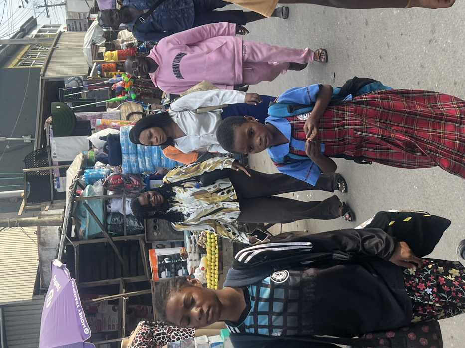The image captures a busy daytime street scene in Egan, Nigeria, featuring multiple pedestrians and market stalls. The ground is a light gray paved surface.
On the left, a purple umbrella marked "MONEY TRANSFER" shades a market stall displaying household goods, including plastic basins, packaged items, and an appliance box. A mannequin torso in a leopard print top stands near this stall. Further right, more market stalls line the street, stocked with various items such as cleaning supplies, plastic containers, and other packaged products. Buildings with corrugated metal siding and overhead utility wires are visible in the background.
Six individuals are prominently featured, all walking generally from left to right across the frame.
1. In the immediate foreground on the left, a young person, likely female, wears a black and blue checkered long-sleeved shirt (resembling a Chelsea FC jersey) over floral-patterned pants. She carries a black backpack and a black bag with yellow patterns.
2. Behind and to the right, an adult female with dark braided hair wears a multi-patterned (gold, black, white) long-sleeved blouse and dark trousers. She carries a black shoulder bag and holds a dark beverage bottle and a smartphone.
3. Centered in the mid-ground, a young girl with short dark hair wears a blue short-sleeved top with a white collar over a long red plaid dress. She has a blue backpack and is seen manipulating a small, light-colored object in her hands while looking towards the viewer.
4. To her right, an adult female with straight dark hair wears a shiny white long-sleeved blouse and dark trousers, carrying a light-colored handbag.
5. Further back on the right, an adult male with short hair and eyeglasses wears a pink hooded sweatshirt with "EGAN UMMU" text and matching pink sweatpants. He wears sandals and carries a light-colored shoulder bag.
6. Partially visible on the far right, an adult male wears a dark blue collared shirt and dark trousers, carrying a dark shoulder bag.
The scene depicts active pedestrian movement within a commercial urban setting.