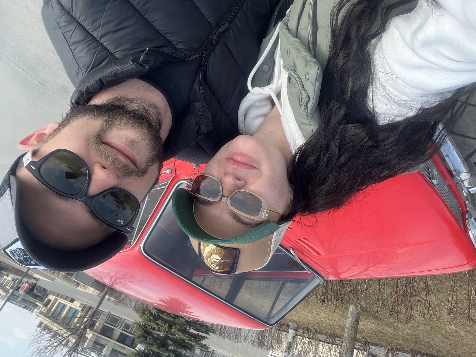 A man and a woman are positioned closely together, seemingly lying on top of a red vintage car. The man is wearing sunglasses and a baseball cap, along with a black puffy jacket. He has a beard. The woman is also wearing sunglasses and a baseball cap with a patch on it. She has long, dark hair. She is wearing a white hoodie and a green jacket.
The red car is clearly visible beneath them, filling much of the frame. The surrounding environment includes trees and buildings in the distance. Based on visual cues, it is not possible to determine the exact location in Kazan, Russia.