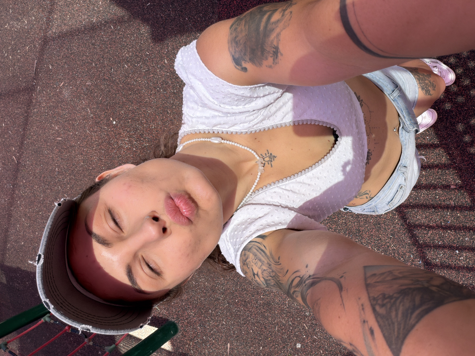 A woman is lying on the ground, apparently on a playground surface, as inferred from the red rubber ground cover and colorful play equipment visible in the periphery. She is oriented at an angle towards the camera, suggesting she is taking a selfie. The woman wears a white top with a beaded necklace, denim shorts, and pink shoes. She has several tattoos on her arms and torso. She has her eyes closed and her lips pursed. The location is Kiryat Motzkin, Israel, based on available location data.