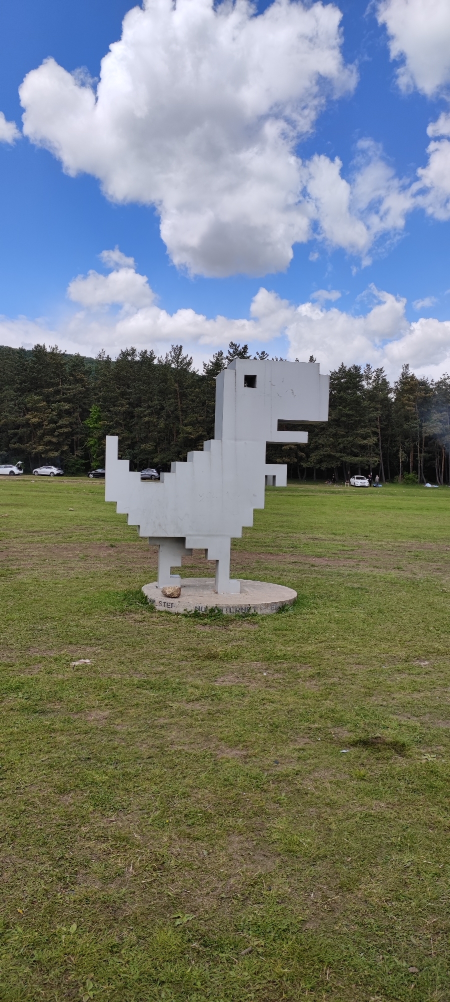 A full shot captures a large, gray, blocky dinosaur sculpture in a grassy field, against a backdrop of trees and a partly cloudy sky. The sculpture, resembling a retro 8-bit design, stands on a circular concrete base. The base has some graffiti/writing on it. In the background, several vehicles are parked, and people are scattered amongst the trees. The scene is located in Gyulagarak, Armenia.