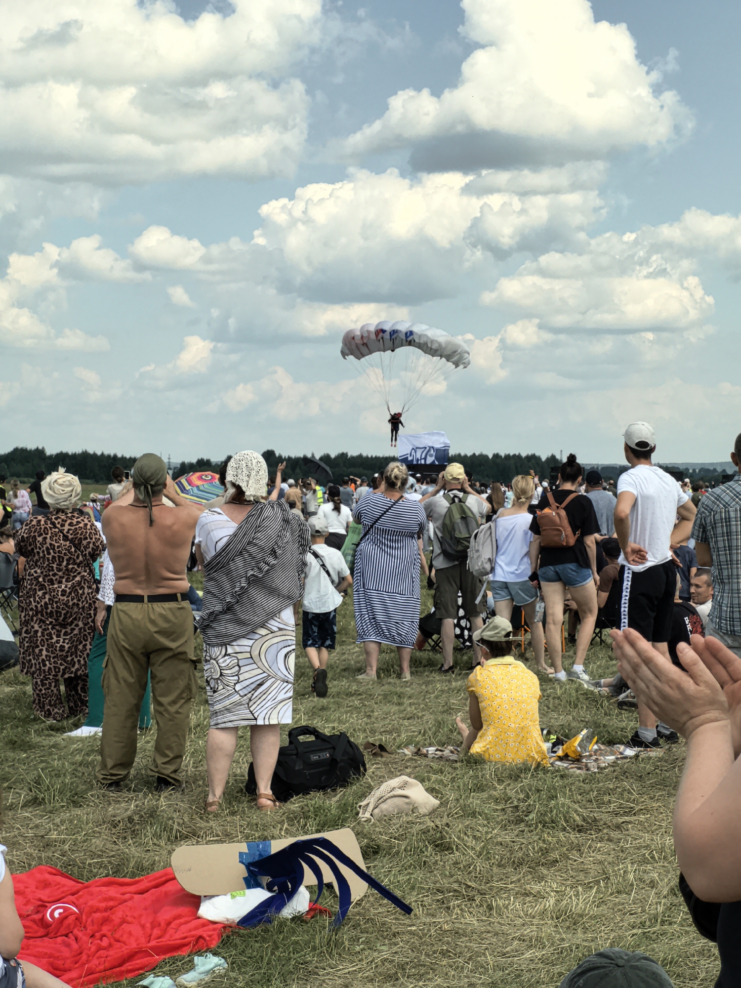 This image depicts an outdoor event likely taking place at an airfield or open field near Nestiukovo, Russia. In the sky, a parachute is visible, descending with a person attached. Below, a crowd of spectators stands on grassy ground, facing the sky and observing the parachute. The crowd consists of individuals of various ages and genders, dressed in casual attire suitable for warm weather, with some wearing hats and headscarves. Several people are holding hands or interacting with each other. Objects scattered on the ground include blankets, bags, and what appear to be picnic items. The environment is characterized by a large, open field under a cloudy sky, with a line of trees visible in the distance. The overall scene suggests a public event, possibly an airshow or festival, where skydiving is being demonstrated.