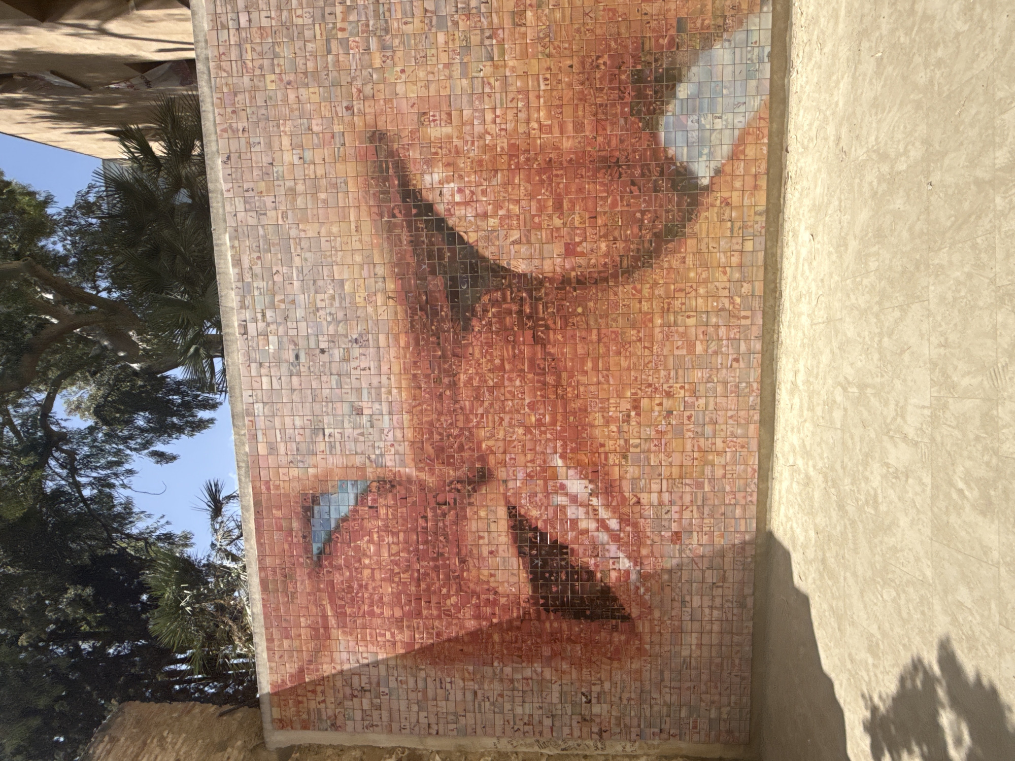 The image displays a large mosaic mural on a textured concrete wall. The mural depicts a close-up of a person's face, rendered in shades of red, orange, and blue tiles. The face is detailed, showing an open mouth, teeth, and an eye. The mural is composed of numerous small, square tiles, each featuring a tiny image. The wall itself is bordered by a thin concrete ledge at the top. Above the wall, there are trees with dark green foliage and a clear blue sky. To the right of the mosaic, a building with a beige facade and a tiled roof is partially visible, casting shadows onto the wall. In front of the wall, the ground is covered in a light-colored, textured surface, possibly pavement or compacted earth, with shadows of trees cast upon it. This artwork is located at the Parc del Carmel in Barcelona, Spain, a public park known for its panoramic city views and unique architectural elements.