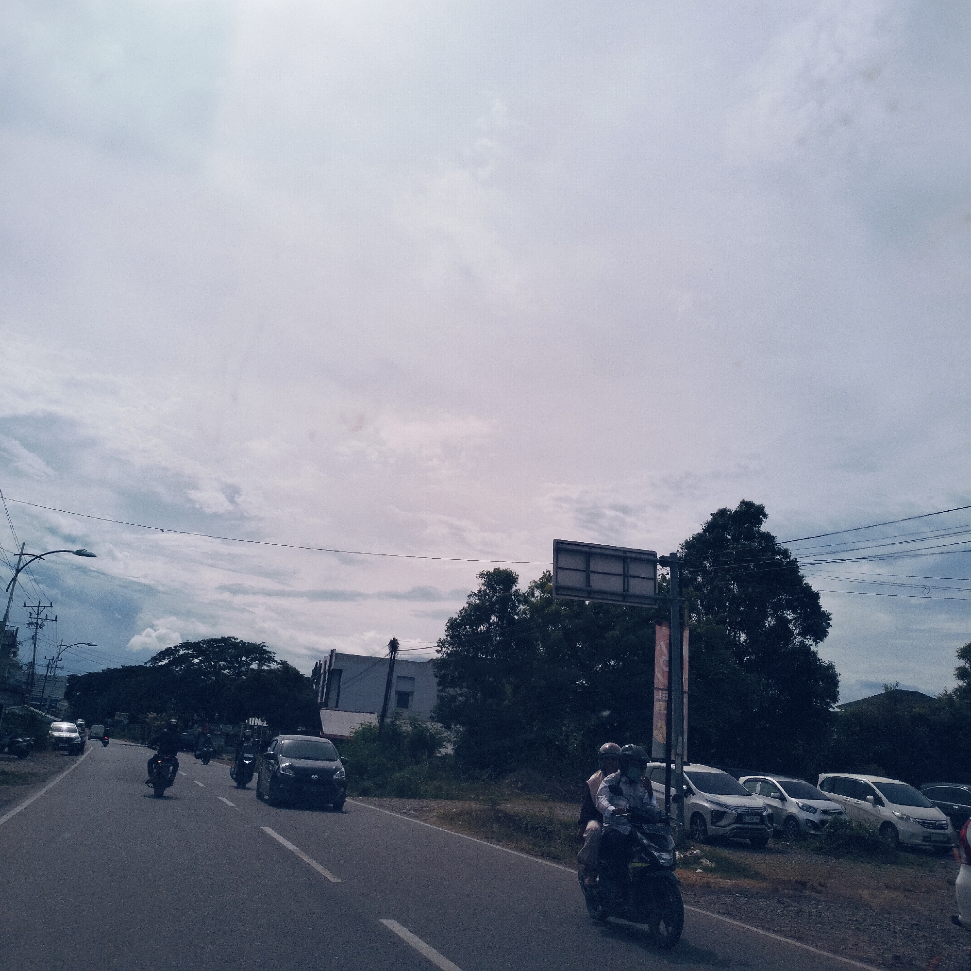 The image depicts a bustling street scene in Cot Teungoh, Indonesia, on an overcast day with a bright, diffused sky. An asphalt road with white lane markings dominates the foreground and mid-ground, showing active traffic. Several motorcycles are visible, with one in the mid-ground carrying two riders, both wearing helmets. One rider is dressed in a light-colored shirt. Further down the road, more motorcycles and cars are in motion or stopped.
On the right side of the road, several cars are parked, including a dark SUV and multiple white vehicles, one resembling a minivan. The roadside features sparse, dry grass and dense, dark green trees characteristic of tropical climates. In the background, light-colored buildings are partially visible through the foliage.
Overhead, utility poles with numerous electrical wires crisscross the sky, alongside a street lamp. A large, blank rectangular sign is mounted on a pole, presumably for advertisements or road information, and another smaller pole with unreadable red and orange banners stands near the parked cars. The scene captures a routine day of travel and activity in a semi-urban setting. No specific emotions are discernible.