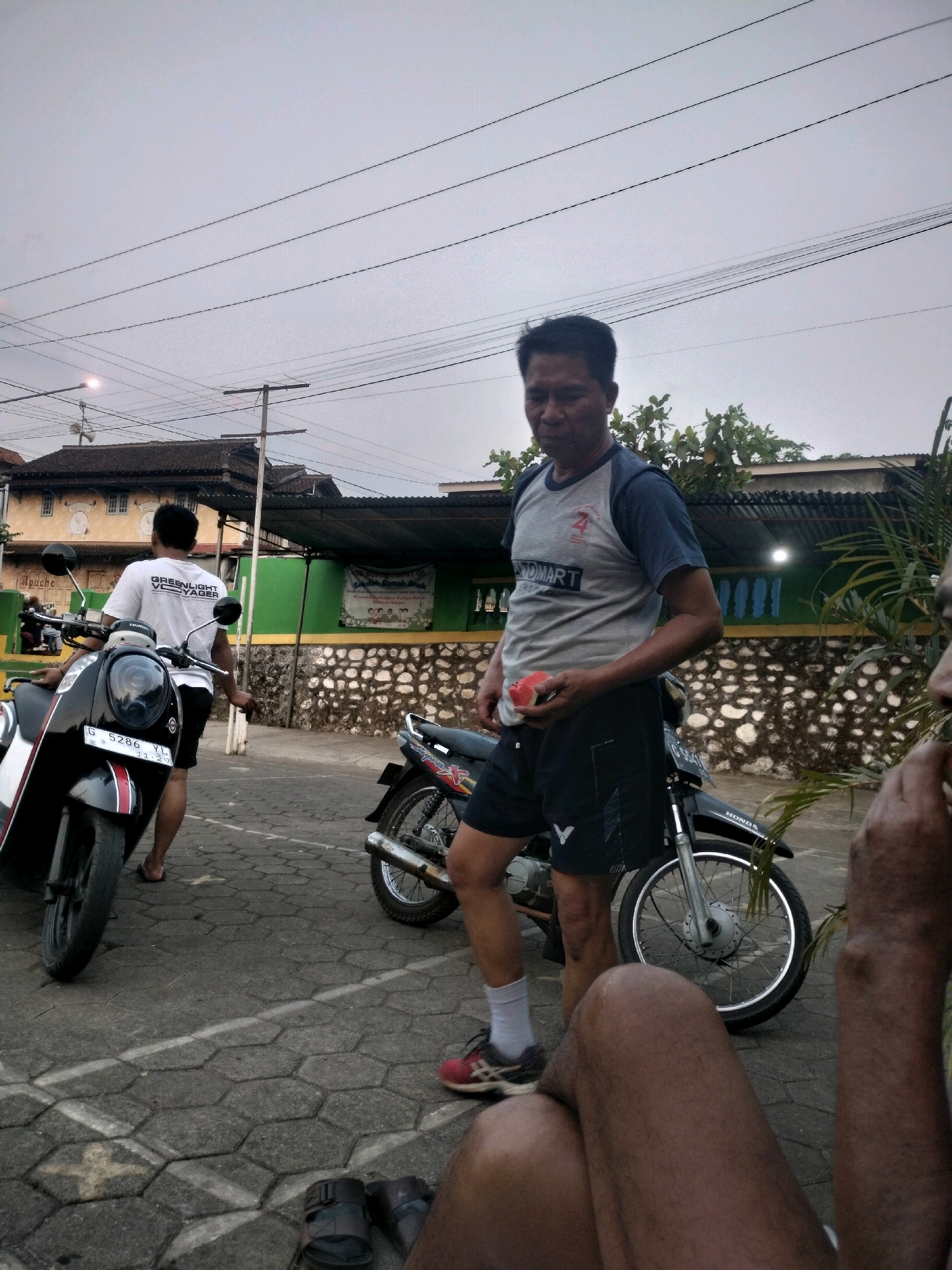 In Boja, Indonesia, a man in a grey and blue t-shirt and black shorts holds a slice of watermelon. He stands next to a motorcycle. Another man in a white t-shirt and black shorts is walking away from a black motorcycle. Both men appear to be in an outdoor setting with buildings and trees visible in the background. The ground is paved with hexagonal stones. The sky is overcast.