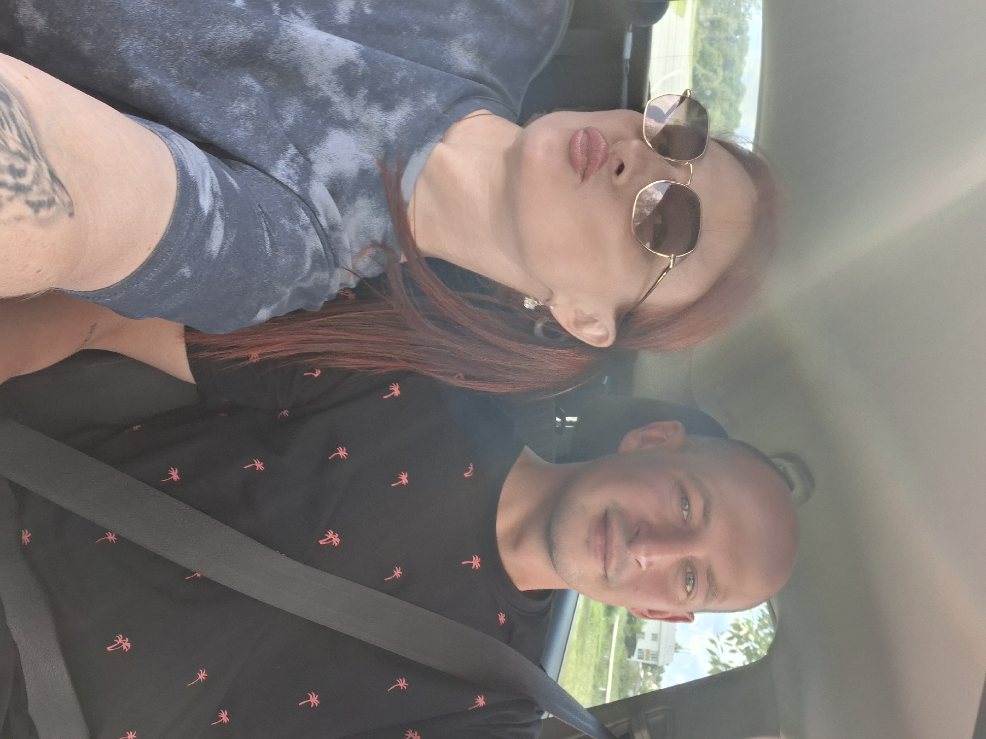 A medium shot captures a man and a woman in the front seats of a car, seemingly on a road trip. The woman, on the left, wears oversized sunglasses and a tie-dye t-shirt, her lips pursed in a playful pout. She has red hair tied back in a ponytail, and a dangling earring is visible. The man, on the right, is bald and wears a black t-shirt with a repeating palm tree pattern. Both individuals are wearing seatbelts, indicating they are traveling. The background shows a blurry view of trees and a building through the car windows, suggesting an outdoor setting, possibly on the outskirts of a town like Myadzyel, Belarus, given the context of the road trip. The overall lighting suggests it is daytime.
