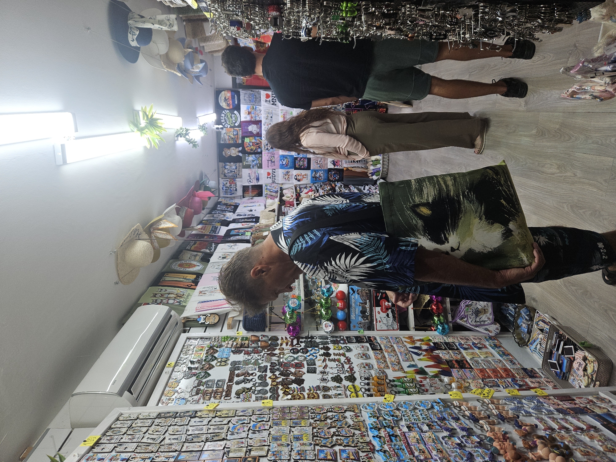 In a souvenir shop in Unknown, Unknown, a man wearing a dark blue and black patterned short-sleeved shirt is browsing items on a wall display. He is holding a large tote bag with a cat graphic on it. Next to him, a woman with long brown hair is wearing a light pink long-sleeved top and olive green pants. Further down the aisle, a man in a black t-shirt and dark green shorts stands with his back to the camera. The shop is filled with various souvenirs, including magnets, keychains, t-shirts, and hats. An air conditioning unit is mounted high on the wall above the man. The floor is light wood laminate.