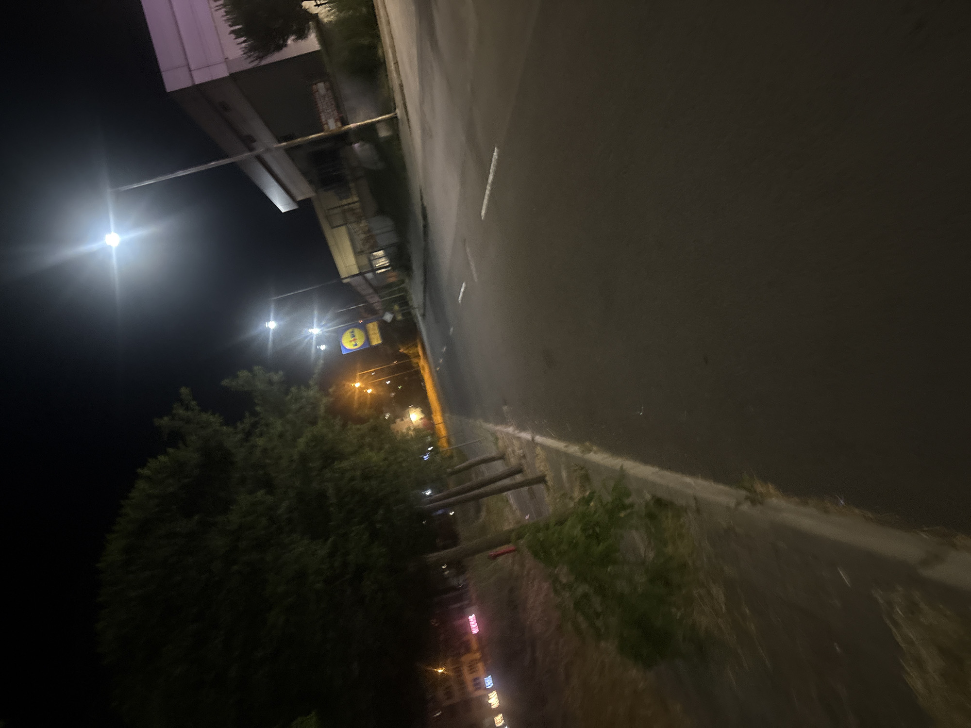 An outdoor, nighttime, low-angle, wide shot captures a street scene in Burgas, Bulgaria. A dark asphalt road stretches into the distance, marked with dashed white lines. On the left side of the road, a row of trees with dark green foliage lines a sidewalk. Beyond the sidewalk, there are some low-lying bushes. Further left, across the street, are some buildings with lights, including what appears to be a Lidl supermarket sign. Streetlights illuminate the scene, casting a bright glow from above. The sky is dark, indicating night. The overall impression is that of an urban street at night, possibly in a commercial or residential area.