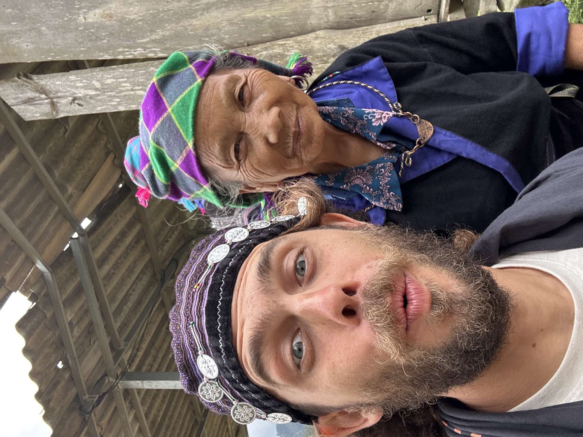 The image displays a close-up selfie featuring an adult male in the foreground and an older female partially visible behind him, set in Dế Xu Phình, Vietnam. The male, positioned to the left, has light brown curly hair, a full beard, and a mustache. He wears a dark, possibly purple, patterned headpiece adorned with multiple round, silver-colored medallions featuring a floral design, a white t-shirt, and a dark outer garment. His eyes are wide open, and his mouth is pursed into an "o" shape, indicating a playful expression as he looks directly at the camera. The female, situated behind and to the right of the male, presents an aged appearance with noticeable facial wrinkles and a subtle smile. She wears a vibrant plaid head covering in green, purple, and grey, with strands of grey hair visible. Her attire includes a dark blue collared shirt featuring a teal and pink floral pattern, layered over a black long-sleeved garment. She accessorizes with a copper-toned necklace holding a small pendant and simple silver hoop earrings. Both individuals are looking towards the camera. The background reveals a rustic, semi-open-air structure with a weathered, corrugated roof supported by metal beams and rough, unpainted wooden planks or posts on the right side.