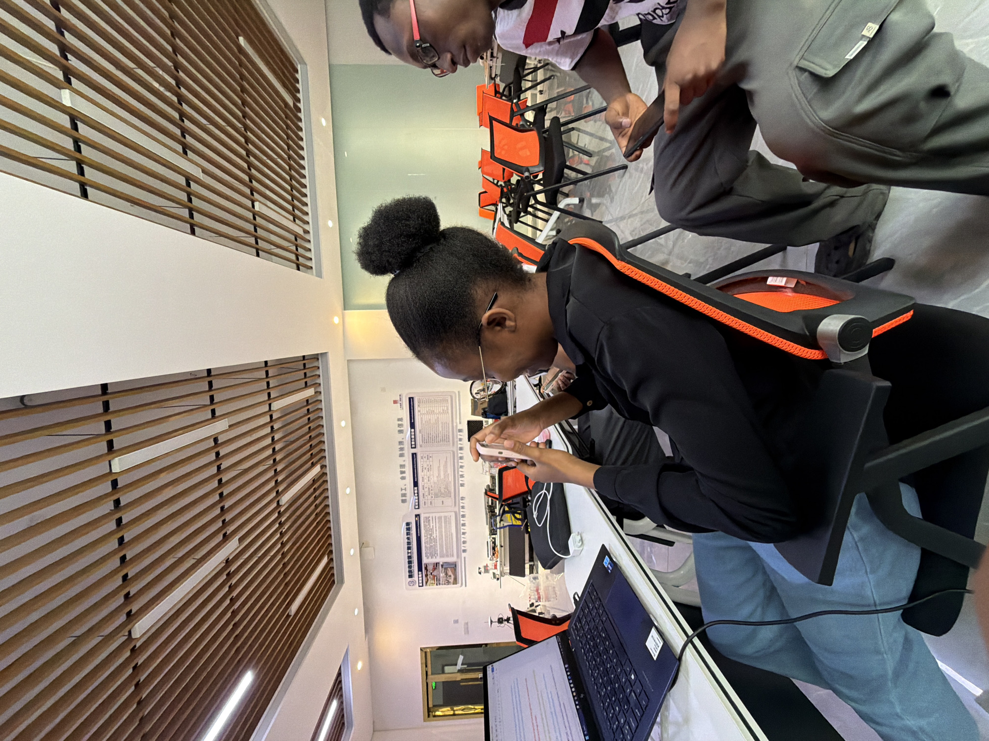 The image depicts an indoor scene, likely an office or classroom, in Jiangjin, China. Two individuals, a woman and a man, are seated at a table. The woman, in the foreground and to the left, is wearing glasses and has her hair in a bun. She is holding a pink smartphone and appears to be looking at it. A laptop is open on the table in front of her, displaying text. The man, to the right, is wearing glasses and a t-shirt with red and black stripes. He is also looking at a smartphone. The room has a modern ceiling with wooden slats and embedded lights. Orange chairs with black frames are visible in the background, suggesting a collaborative or study environment. A poster with Chinese text is mounted on the wall behind the individuals. The overall impression is one of a contemporary workspace or educational setting.