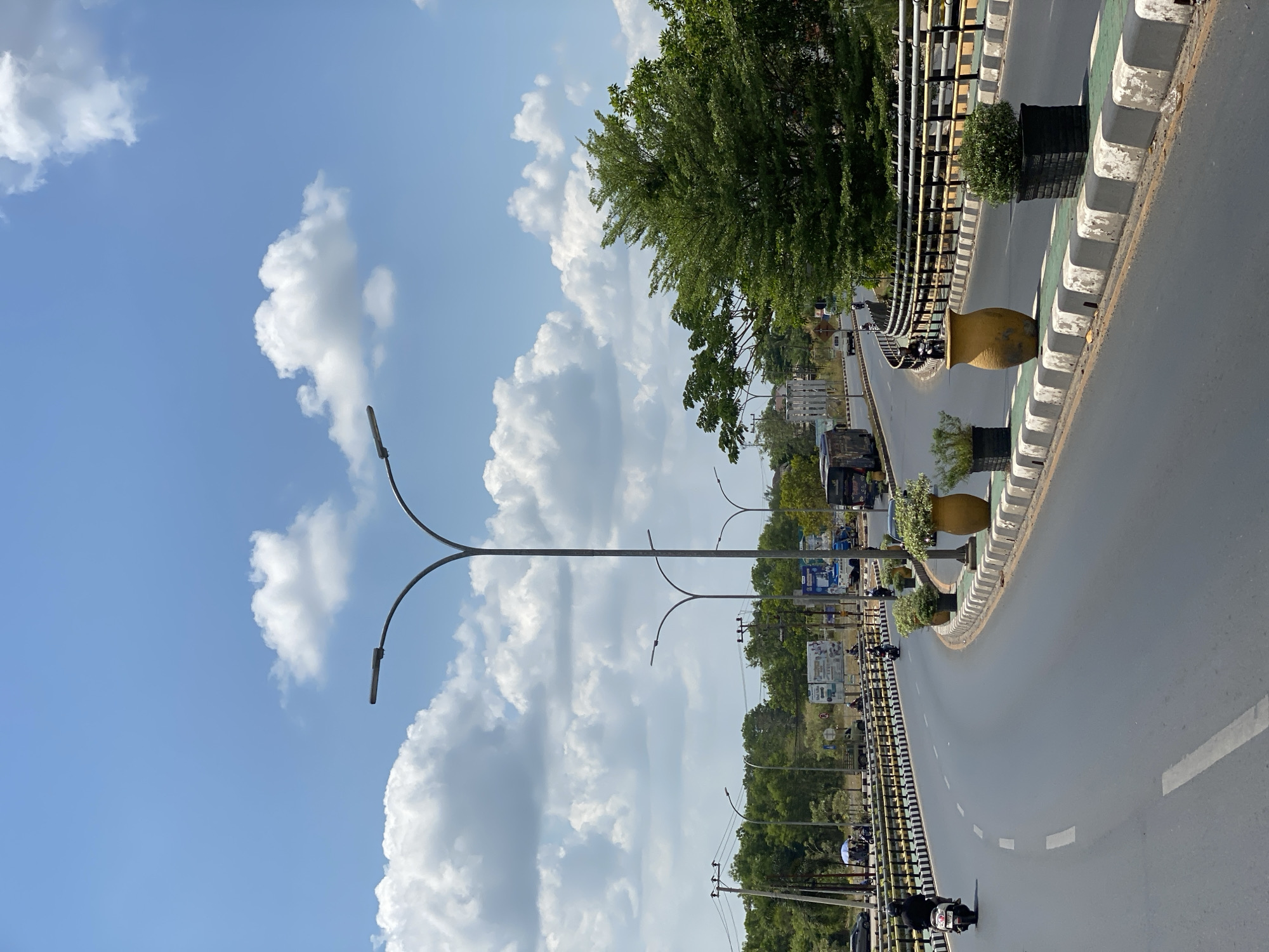 The image shows a bright, sunny day in Banjarmasin, Indonesia, with a wide, curving road dominating the foreground. The road has a smooth asphalt surface with clear white lane markings. Along the sides of the road are decorative planters containing green plants and a large, yellow vase. A black metal railing with yellow accents separates the road from the embankment. In the background, several streetlights with modern, sleek designs line the road. Various vehicles, including motorcycles and cars, are visible on the road and in the distance. Trees and buildings can be seen beyond the road, suggesting an urban or suburban environment. The sky is a vibrant blue with scattered fluffy white clouds. The overall scene conveys a sense of an active, well-maintained urban infrastructure.