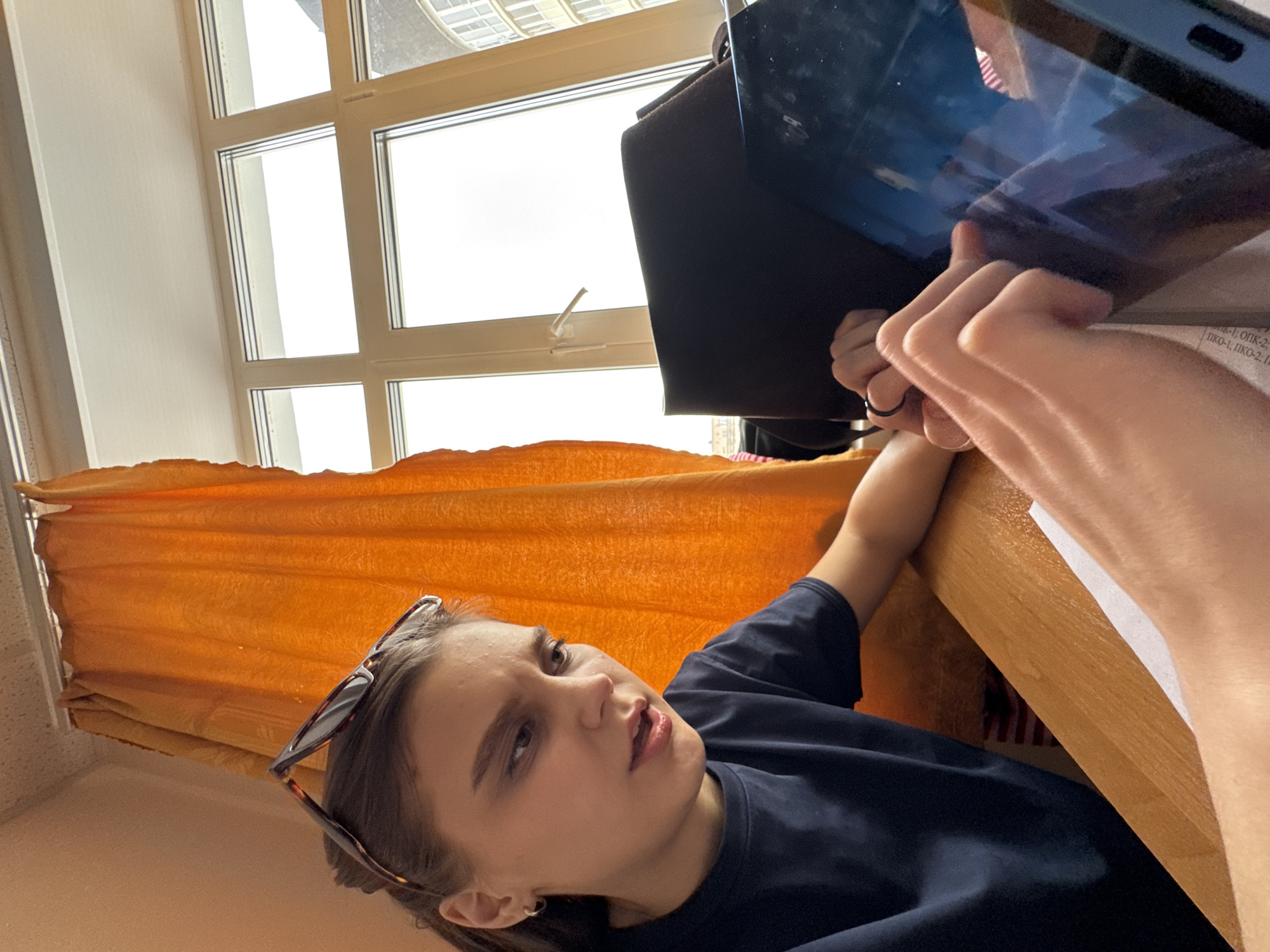 The image depicts an indoor scene in Perm, Russia, focusing on a young female seated at a light-colored wooden desk. She wears a dark blue short-sleeved t-shirt, and tortoiseshell sunglasses are positioned on top of her dark hair. Her left arm rests on the desk, while her right hand actively interacts with the screen of a blue-cased digital tablet. The tablet's dark screen reflects ambient light and blurred shapes. Behind the tablet, a black bag or object is visible. To the left, an orange curtain hangs, partially obscuring a white-framed, multi-pane window. Through the window, a bright, possibly overcast sky is visible, along with the upper section of a modern building featuring curved architecture on the far right. A white sheet of paper with faint text is present beneath the subject's right hand. The individual's facial expression suggests concentration or slight bewilderment, with her mouth slightly open.
