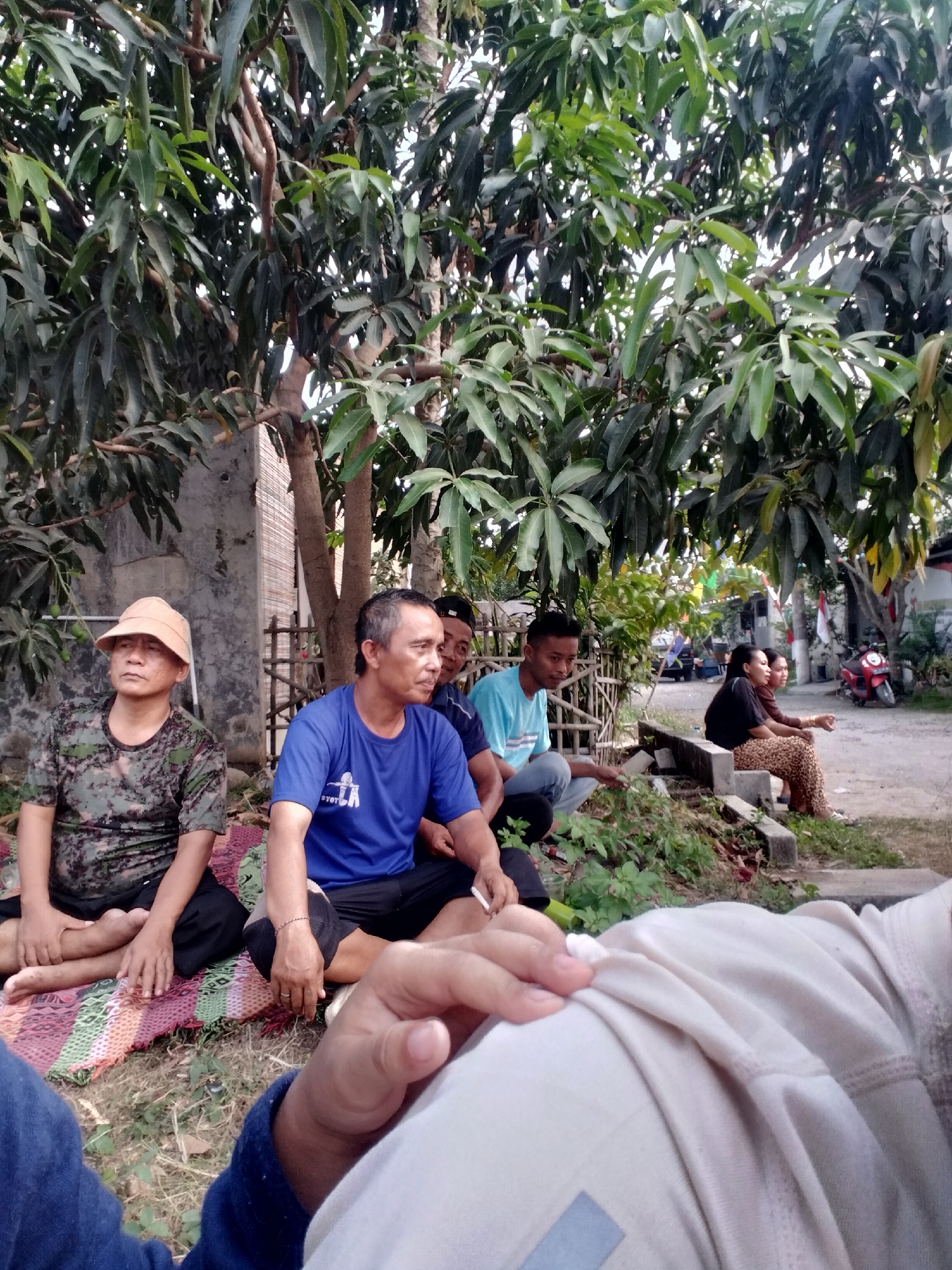 In Cariu, Indonesia, a group of people are gathered outdoors, seated on the ground under a large tree. The foreground is dominated by the back of someone wearing a light-colored long-sleeved shirt. In the middle ground, several men are seated on a colorful woven mat. One man on the left wears a camouflage t-shirt and a light-colored hat. Next to him, a man in a blue t-shirt is holding a cigarette. To his right, two other men are also seated, one in a light blue t-shirt and jeans, and another partially visible behind him. In the background, two women are seated on a concrete ledge. One woman in the foreground is wearing a black top and leopard-print pants, while the woman behind her wears a brown top and pants. A red motorcycle is parked in the background, and various buildings and vegetation are visible, suggesting a residential or community outdoor area. The lighting indicates it is likely daytime, possibly late afternoon.