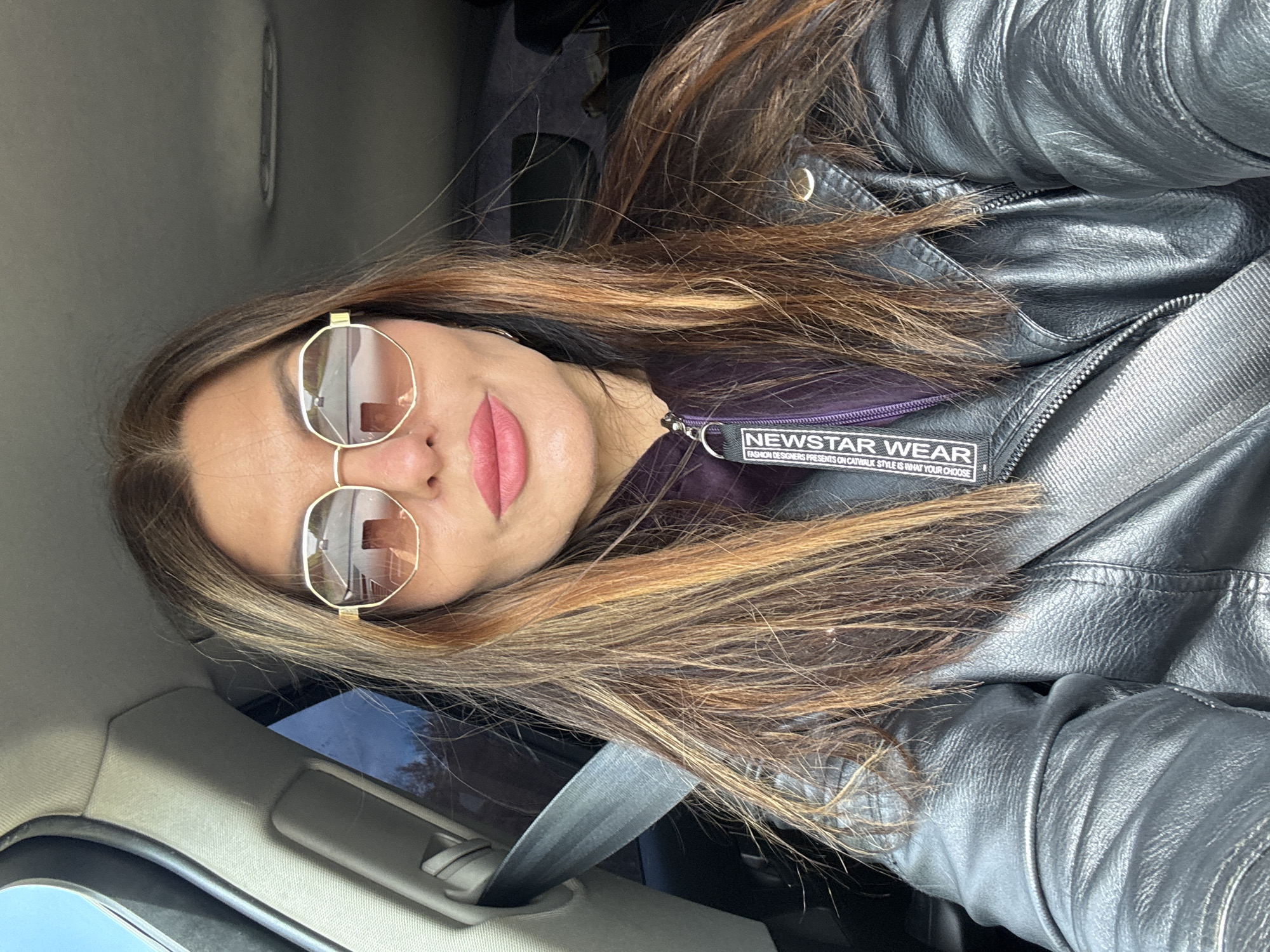 The image shows a woman sitting in a car, wearing a black leather jacket, sunglasses, and a seatbelt. The jacket has a tag that reads "NEWSTAR WEAR." The interior of the car is visible. The location is Chaikovsky, Russia.