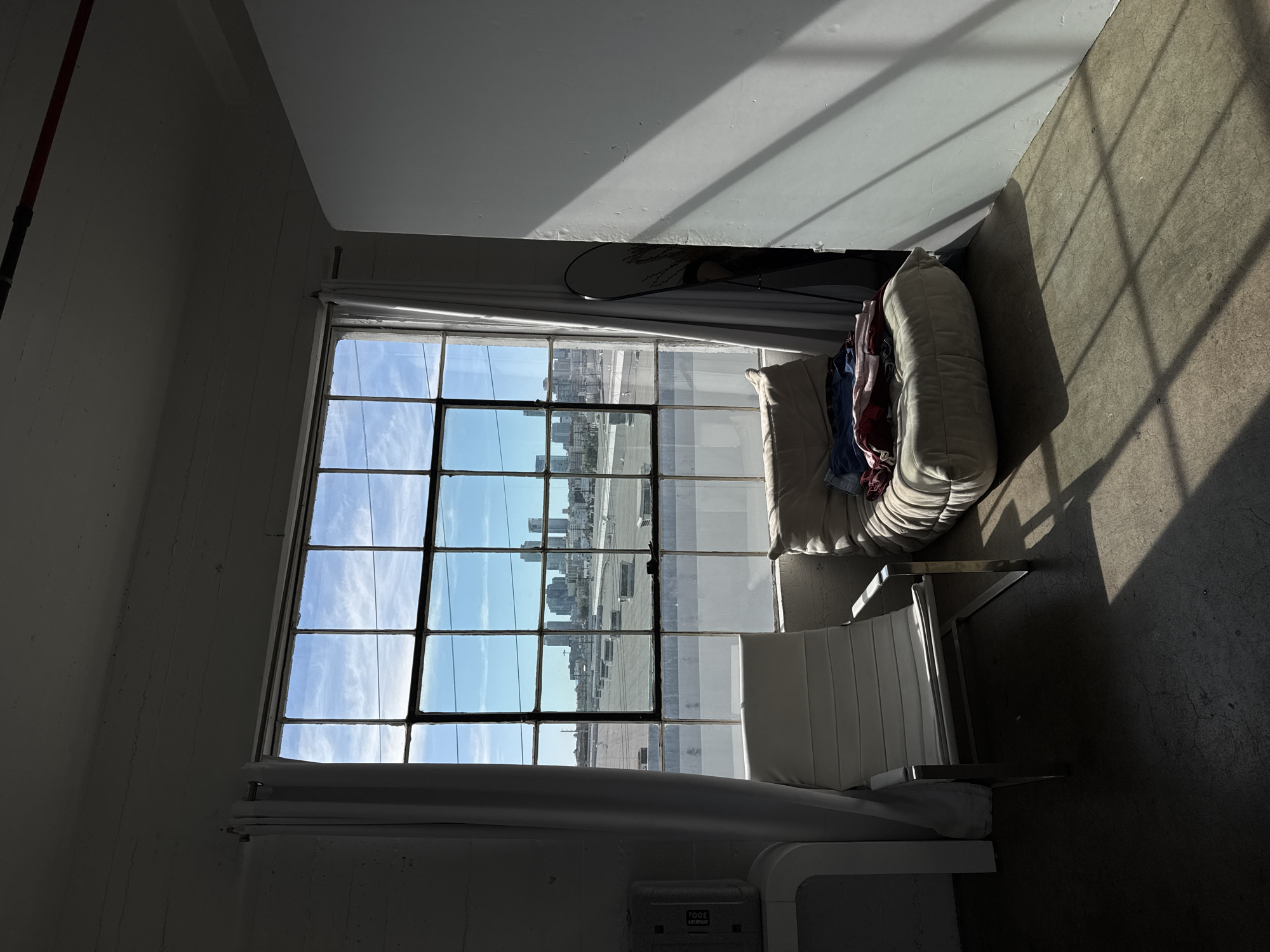 The image is an indoor shot of a room with a large window overlooking a city. The window is divided into multiple panes, and through it, one can see a cityscape with tall buildings, indicative of a major urban area. The sky visible through the window is blue with some clouds. Inside the room, near the window, there's a chair with a white cushion and a modern design. Next to it, there is another soft chair with some clothes in it. The room appears to have white walls. Strong sunlight streams in through the window, creating stark contrasts of light and shadow on the floor and walls. The city visible through the window appears to be Los Angeles, United States of America, with the distinctive downtown skyline.