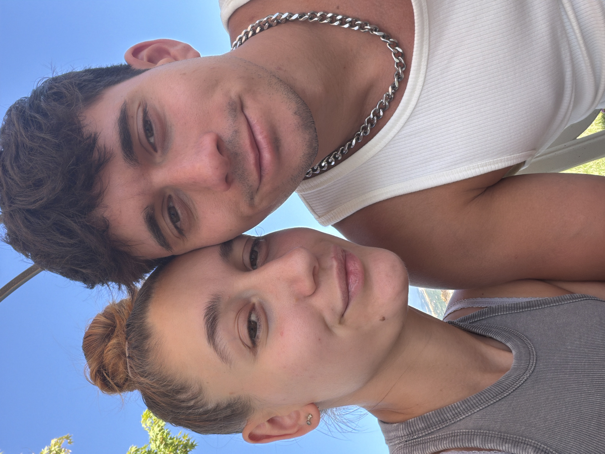 A close-up selfie features a young couple embracing outdoors, with a bright blue sky and hints of greenery in the background. The woman, on the left, has her hair in a bun and is wearing a grey tank top and small earrings. The man, on the right, has dark hair and is wearing a white tank top and a silver chain necklace. They are positioned very close together, with their heads tilted towards each other, suggesting an intimate moment. The environment appears to be sunny and warm, possibly during the daytime. The context of Kocherinovo, Bulgaria is not discernible from the image itself.